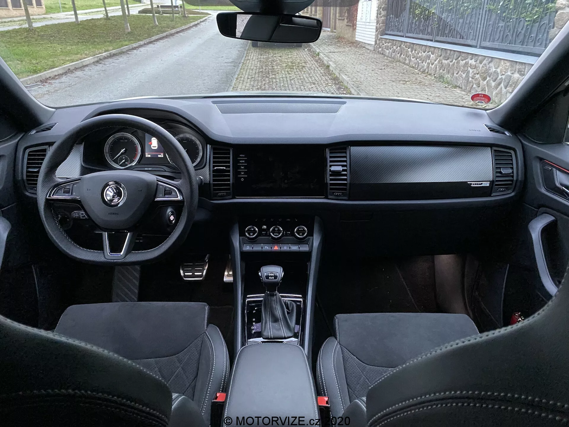 Vue intérieure de la Skoda Kodiaq 2017 depuis l'espace des sièges arrière, montrant le tableau de bord, le volant avec boutons multifonctions, le tableau de bord numérique, l'écran d'info-divertissement large, les commandes HVAC, le sélecteur de vitesse automatique, les matériaux en cuir haut de gamme et les sièges sportifs avec appuie-têtes intégrés.