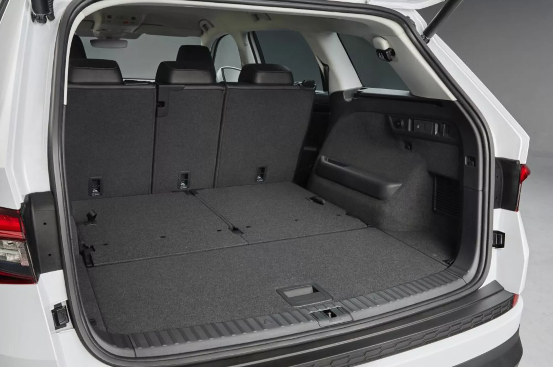 Vue de l'espace de chargement arrière Skoda Kodiaq 2017 avec hayon ouvert, présentant un espace de chargement moquetté vide, sièges arrières rabattables pour un espace élargi, garniture latérale en plastique avec compartiment de rangement encastré, points d'ancrage et éclairage intérieur, soulignant des solutions pratiques de stockage et de gestion du chargement.