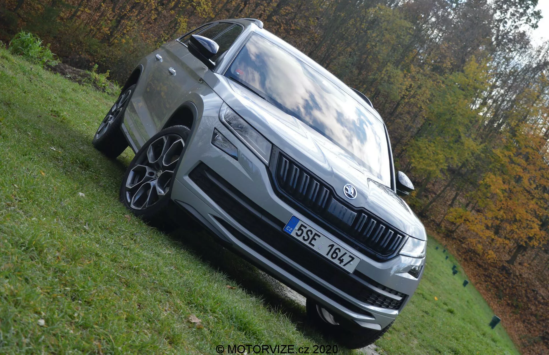 Photographie en contre-plongée d'un Skoda Kodiaq 2017 mettant en valeur sa stature imposante. La voiture est capturée en montrant son quart avant gauche avec des caractéristiques notables telles que des phares à LED, une calandre proéminente avec l'emblème Skoda, des jantes en alliage à plusieurs rayons, un pare-chocs avant avec prises d'air, un rétroviseur avec clignotant intégré et des lignes de design extérieur distinctives. Le véhicule est de couleur grise claire ou argentée et est garé sur une zone herbeuse sous la lumière naturelle du jour. L'arrière-plan flou aux couleurs automnales accentue la présence du véhicule.