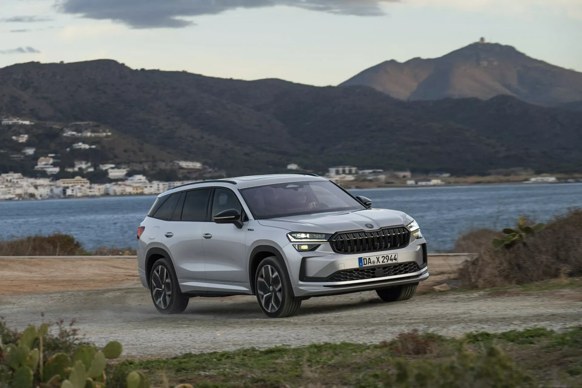 SUV Skoda Kodiaq 2024 garé sur une surface non pavée, présentant un arrière-plan de collines et d'eau, mettant en lumière les phares modernes à LED du véhicule, la grille avant avec intégration ADAS potentielle, et le design contemporain des roues sous une lumière diurne douce et nuageuse.