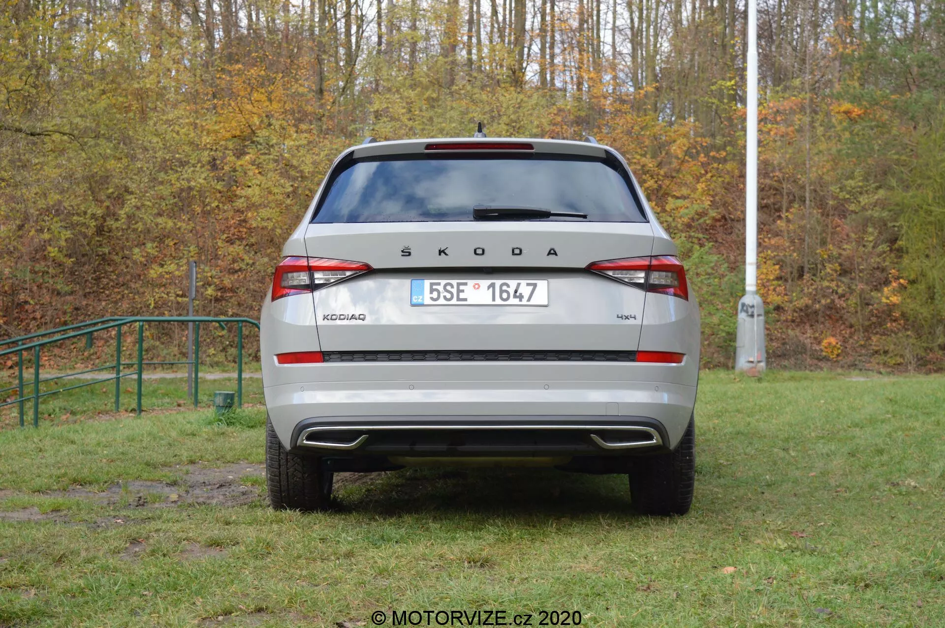 Vue arrière d'un véhicule Škoda Kodiaq 2017 dans un environnement extérieur. La marque 'ŠKODA' et le nom du modèle 'KODIAQ' sont affichés en gros caractères sur le couvercle du coffre. Plaque d'immatriculation de style européen, feux arrière modernes avec feu arrière de brouillard et feux de recul intégrés, pare-chocs arrière visible avec capteurs de stationnement et pare-brise arrière avec essuie-glace sont clairement visibles. Des éléments réfléchissants sur la partie inférieure du pare-chocs améliorent la visibilité. La voiture semble être garée sur une zone herbeuse légèrement boueuse, ce qui suggère une capacité tout-terrain.