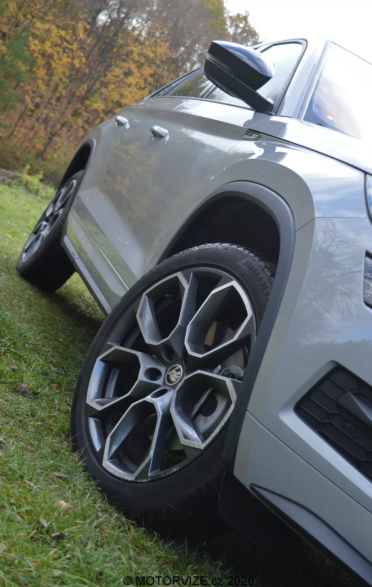 Vue en gros plan et en contre-plongée d'une Škoda Kodiaq 2017 mettant en avant la roue avant et l'aile. La voiture, garée sur l'herbe, présente une roue en alliage à plusieurs rayons, un rétroviseur aérodynamique avec clignotant intégré, des panneaux de carrosserie angulaires avec une finition de peinture métallisée et une prise d'air sur l'aile avant, le tout sur un fond naturel flou.
