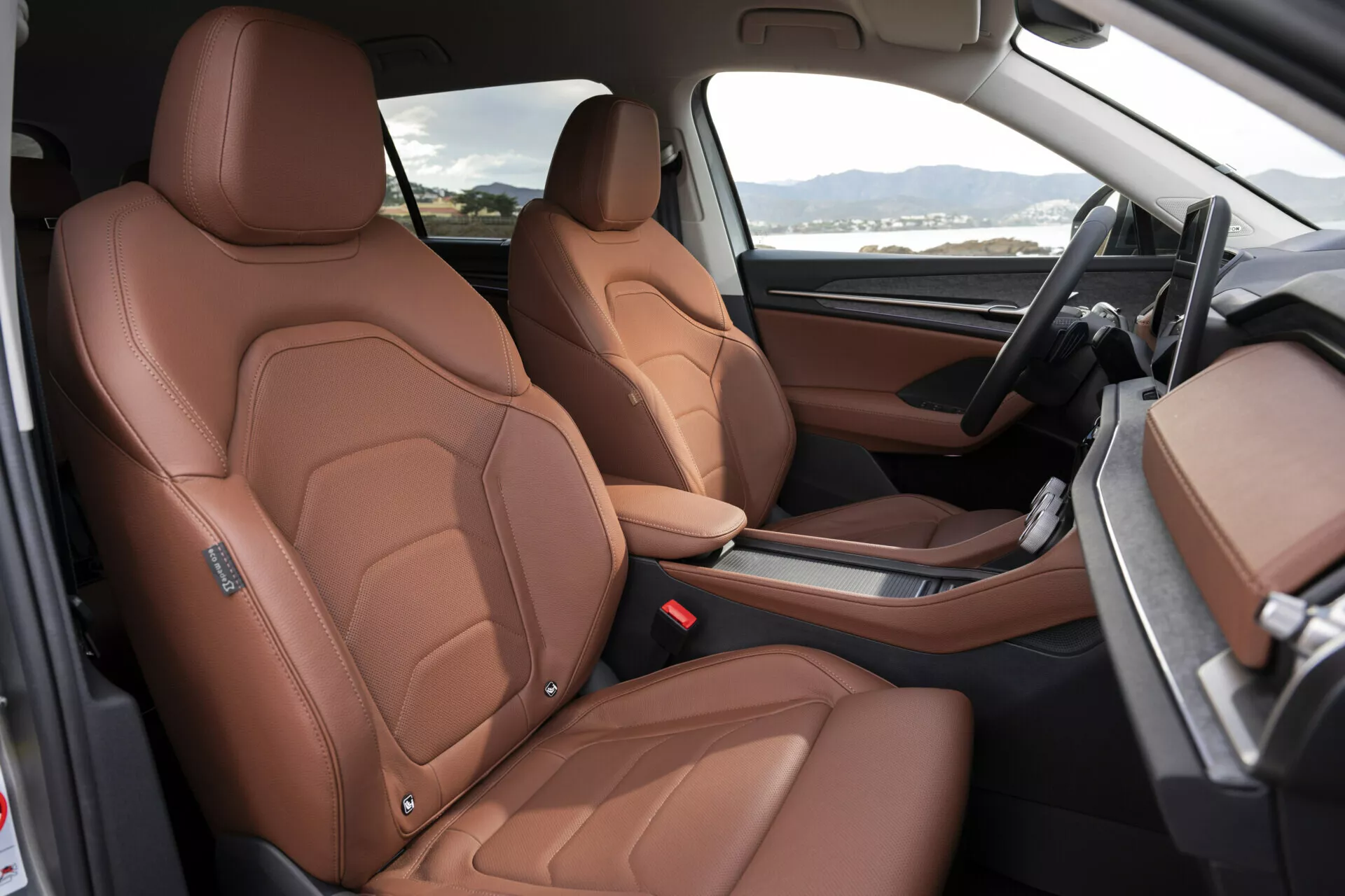 Vue intérieure du Škoda Kodiaq 2024 mettant en avant des sièges avant en cuir marron luxueux avec coutures détaillées, réglage électrique des sièges, et aperçu des éléments de design haut de gamme tels que les renforts latéraux d'inspiration sportive, tableau de bord en deux tons noir et marron, et console centrale minimaliste avec levier de vitesse automatique, depuis une perspective côté passager ouvert
