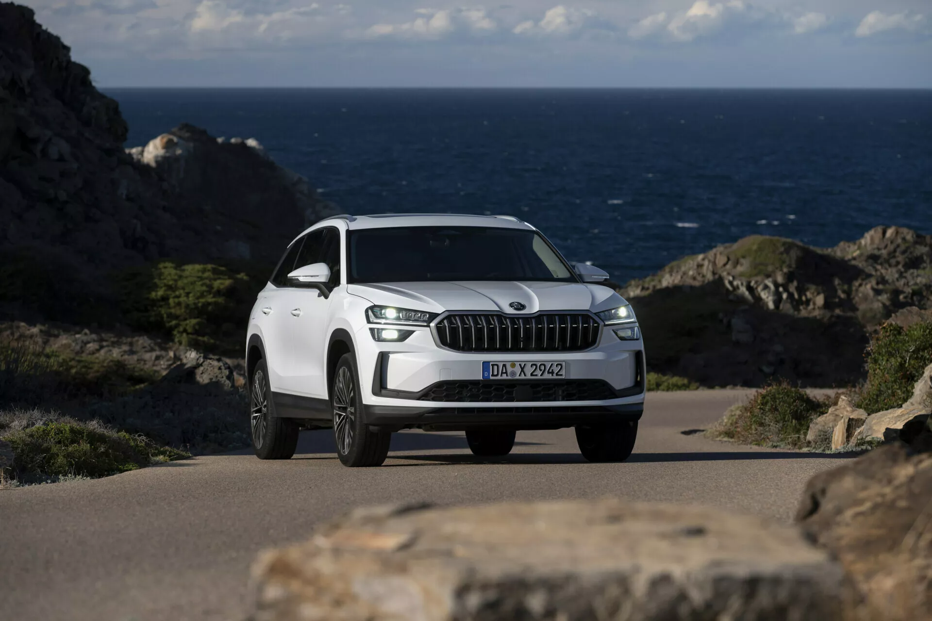 SUV blanc Skoda Kodiaq 2024 garé sur une route côtière avec l'océan en arrière-plan, présentant une grille frontale proéminente avec des lamelles verticales type chrome, des feux de jour à LED, des roues en alliage à multi-rayons et une vue détaillée trois quarts avant