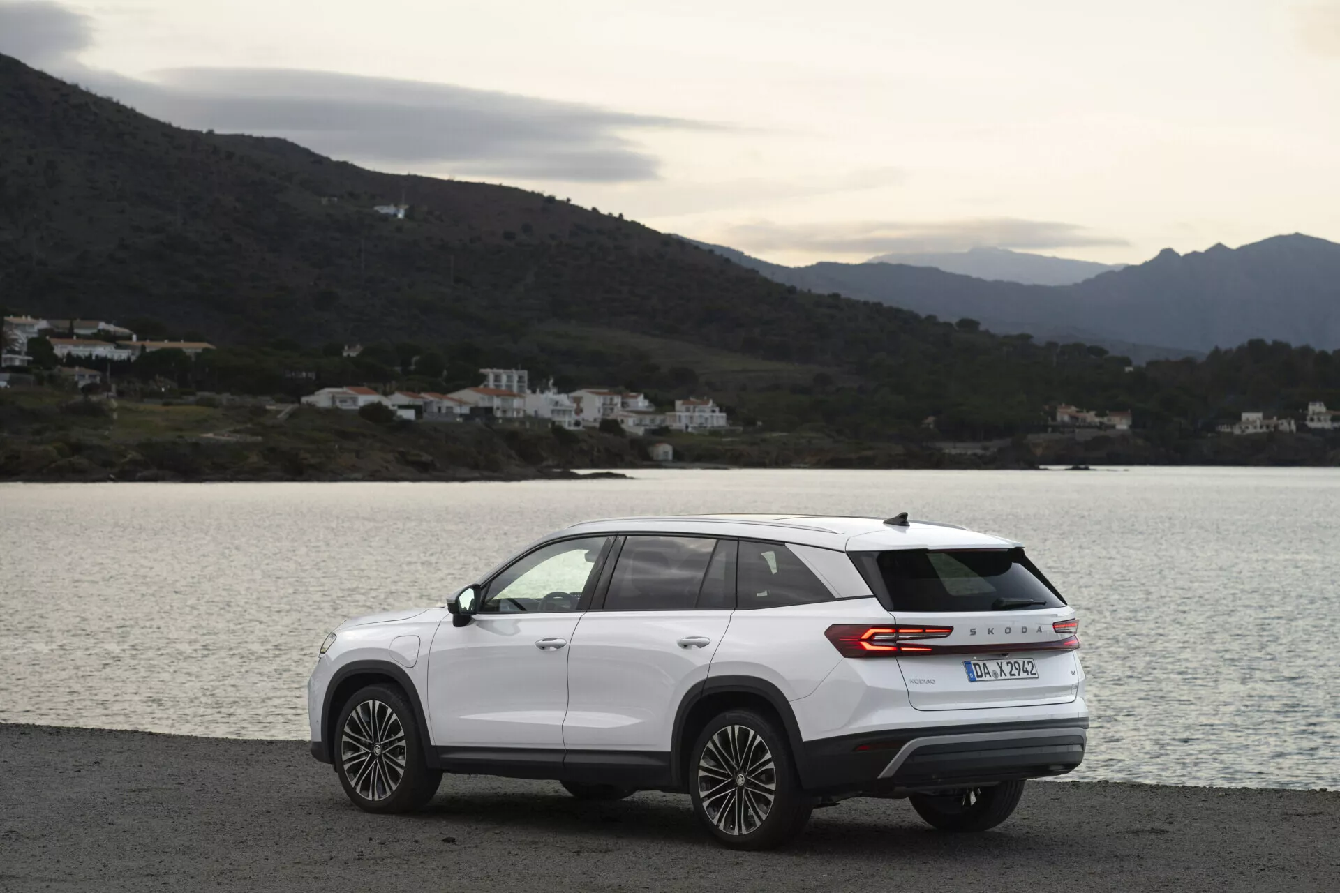 SUV moderne Skoda Kodiaq 2024 garé latéralement sur une route côtière pittoresque, présentant un design aérodynamique, un bandeau lumineux arrière et des jantes en alliage à plusieurs branches, lors d'une journée nuageuse avec un éclairage naturel diffus, sur fond d'eau, de collines et de bâtiments