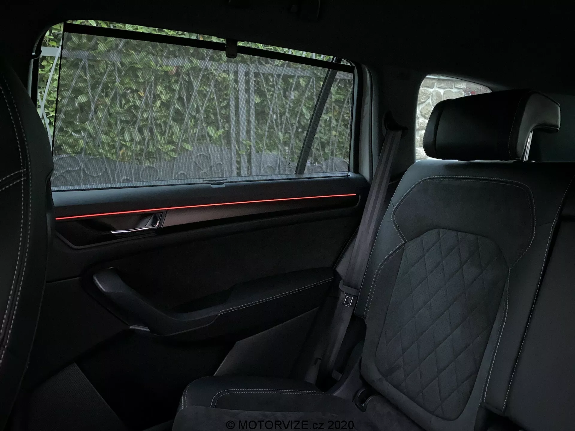 Vue intérieure du Skoda Kodiaq 2017 centrée sur le compartiment arrière des passagers, affichant des caractéristiques de luxe telles que la sellerie en cuir à surpiqûres en losanges sur les sièges avant et arrière, une bande lumineuse ambiante rouge sur le panneau de porte bien équipé avec des garnitures premium, et un pare-soleil partiellement transparent sur la fenêtre. L'accent est mis sur le confort et des éléments de design haut de gamme.