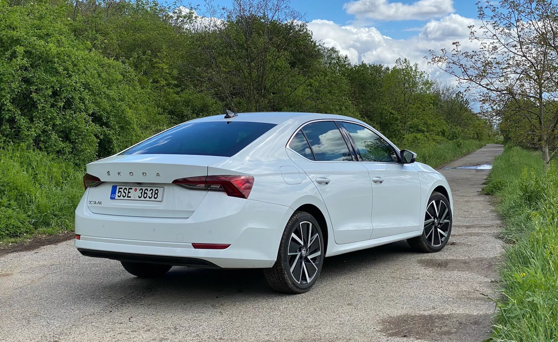 Vue trois quarts arrière d'une berline blanche Skoda Octavia 2020 garée sur le côté droit d'une route étroite, montrant le côté passager depuis le niveau du sol. La voiture remplit la majeure partie du cadre, mettant en valeur les lignes de design, les feux arrière enveloppant le coffre, le subtil spoiler intégré, le badge arrière, les jantes en alliage multi-branches, la vitre arrière légèrement teintée et les rétroviseurs latéraux avec clignotants intégrés.