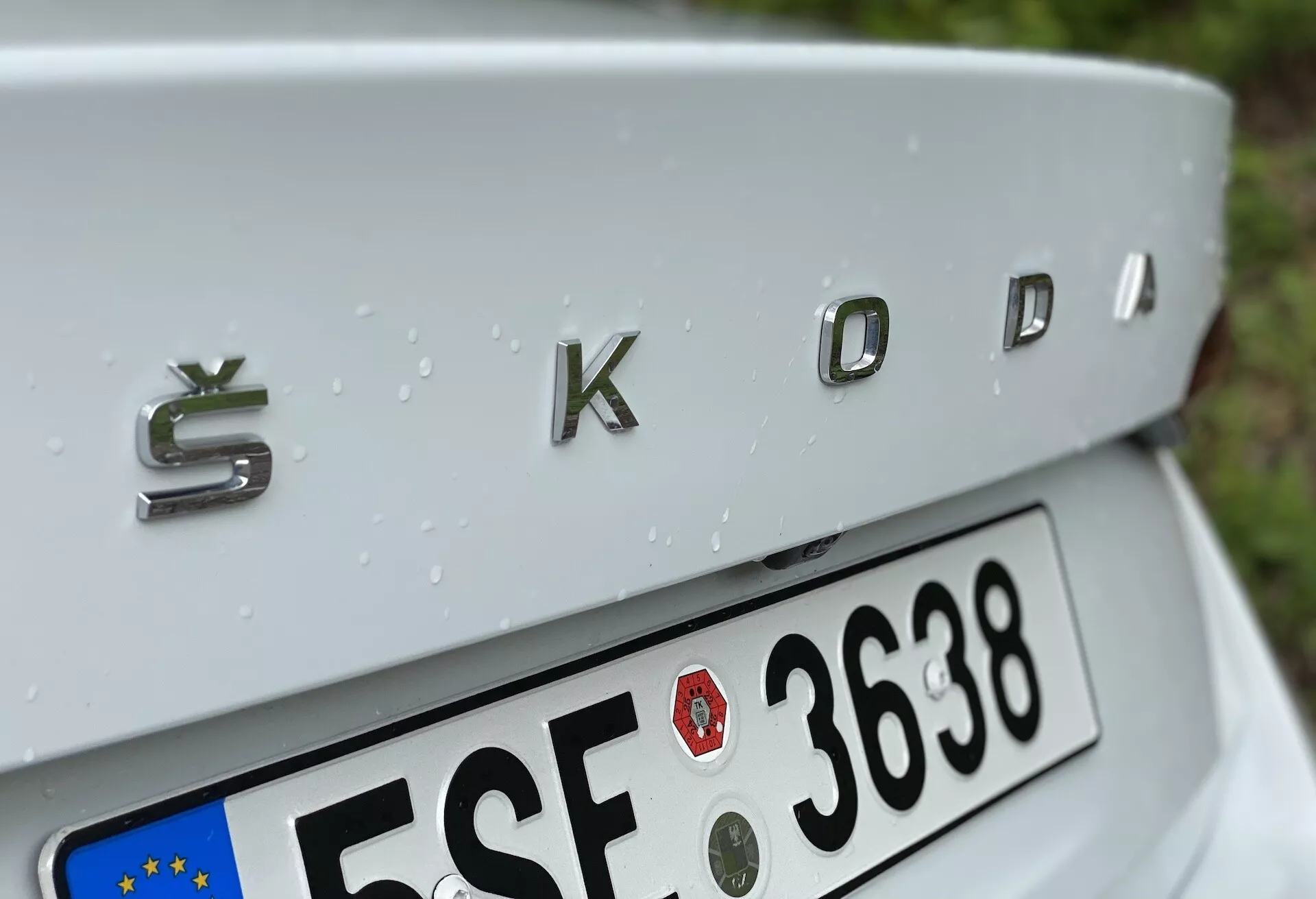 Gros plan de la section arrière d'une Škoda Octavia 2020 (octavia-limousine) centrée sur le badge chromé 'ŠKODA' avec une finition métallique tridimensionnelle. L'angle de prise de vue légèrement en plongée montre le couvercle du coffre doucement incliné, une plaque d'immatriculation de style Union Européenne partiellement visible en dessous et des gouttes de pluie ornant la surface argentée de la voiture, indiquant une pluie récente.