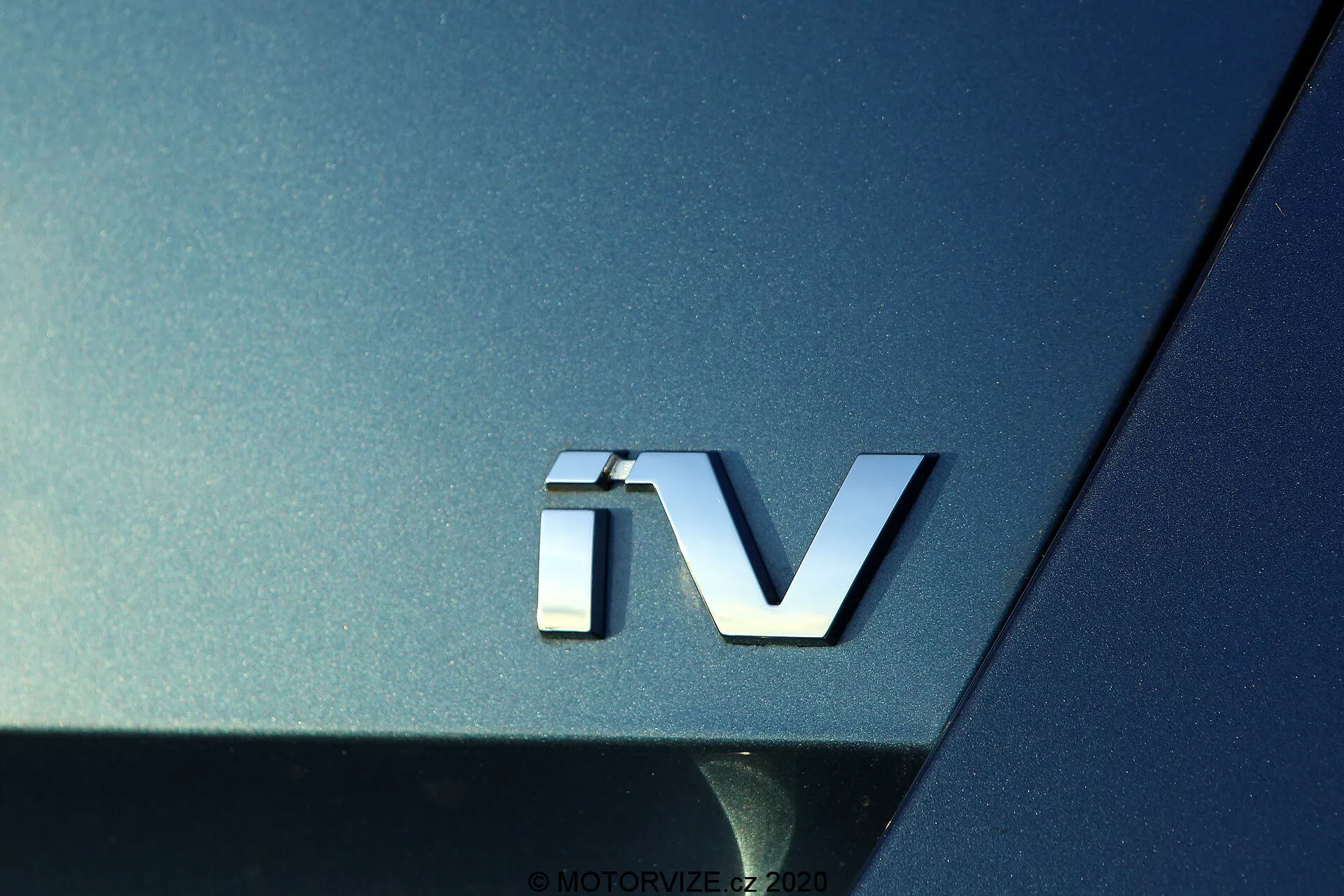 Gros plan sur le badge 'iV' de la Skoda Octavia 2020, indiquant une motorisation électrifiée, avec un emblème argenté en 3D sur la carrosserie métallique sombre, soulignant la surface incurvée et les qualités réfléchissantes de la section arrière de la voiture.