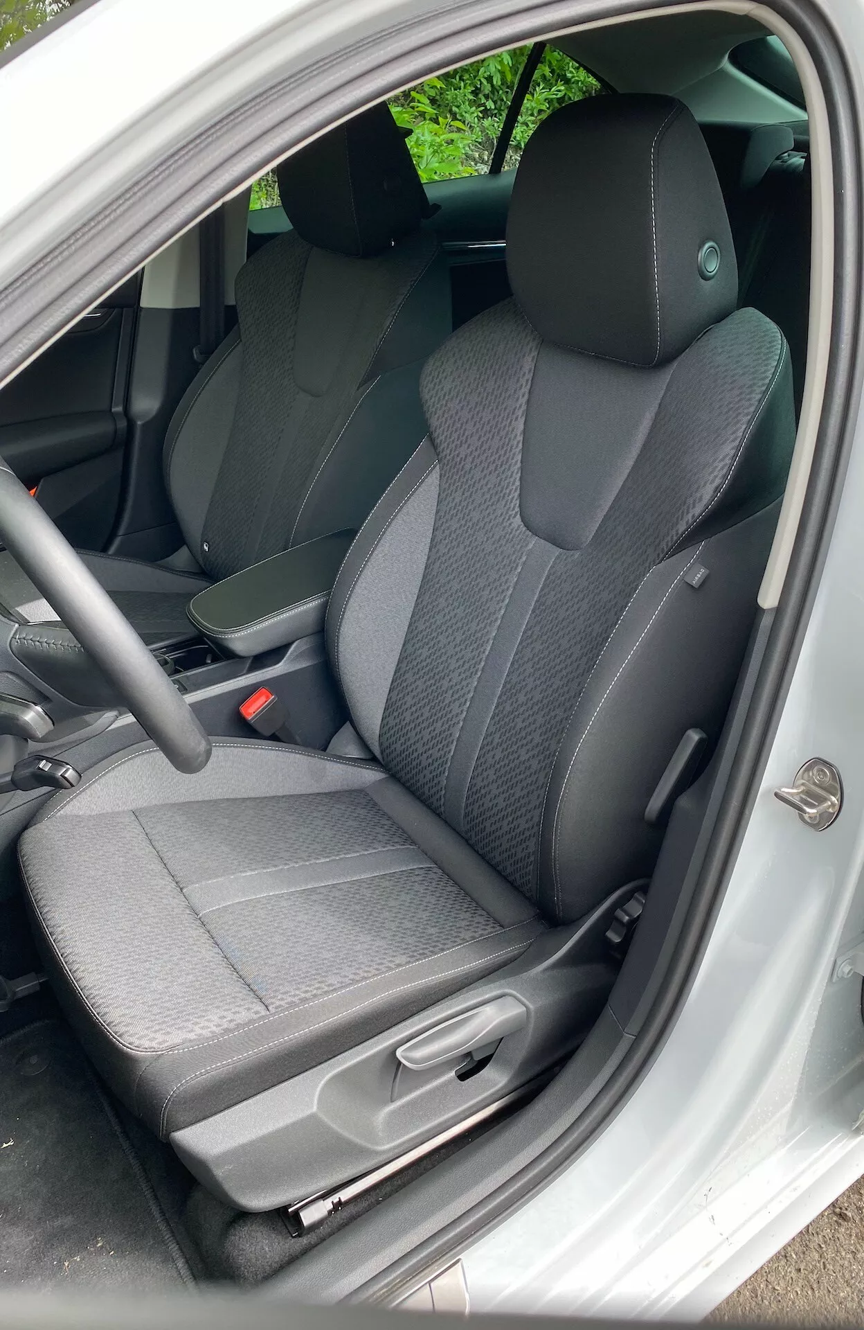 Intérieur de la Škoda Octavia Limousine 2020 vu du côté conducteur avec la porte ouverte. Comprend un siège en tissu bicolore, un tableau de bord simple, un volant standard, un tapis de sol sombre, un réglage manuel du siège et un design intérieur basique.