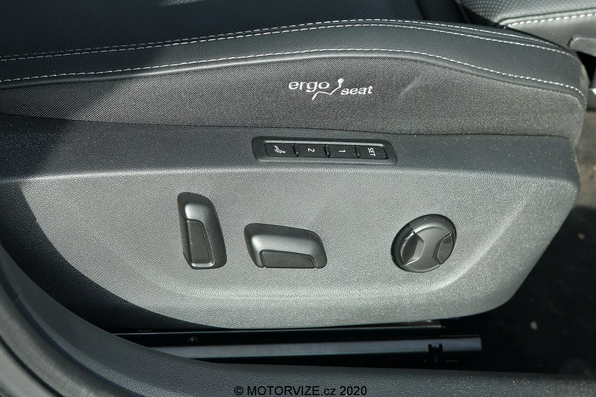 Image en gros plan montrant les commandes du système de siège ergo de la Škoda Octavia Limousine 2020. La photo met en évidence plusieurs boutons et un bouton rotatif pour régler la position, la hauteur, l'inclinaison et le support lombaire du siège du conducteur ou du passager. Le texte 'ergo seat' est visiblement brodé au-dessus des commandes, mettant l'accent sur le design ergonomique. Le siège est en tissu tissé gris foncé, suggérant un matériau durable et confortable adapté aux véhicules de milieu à haut de gamme.