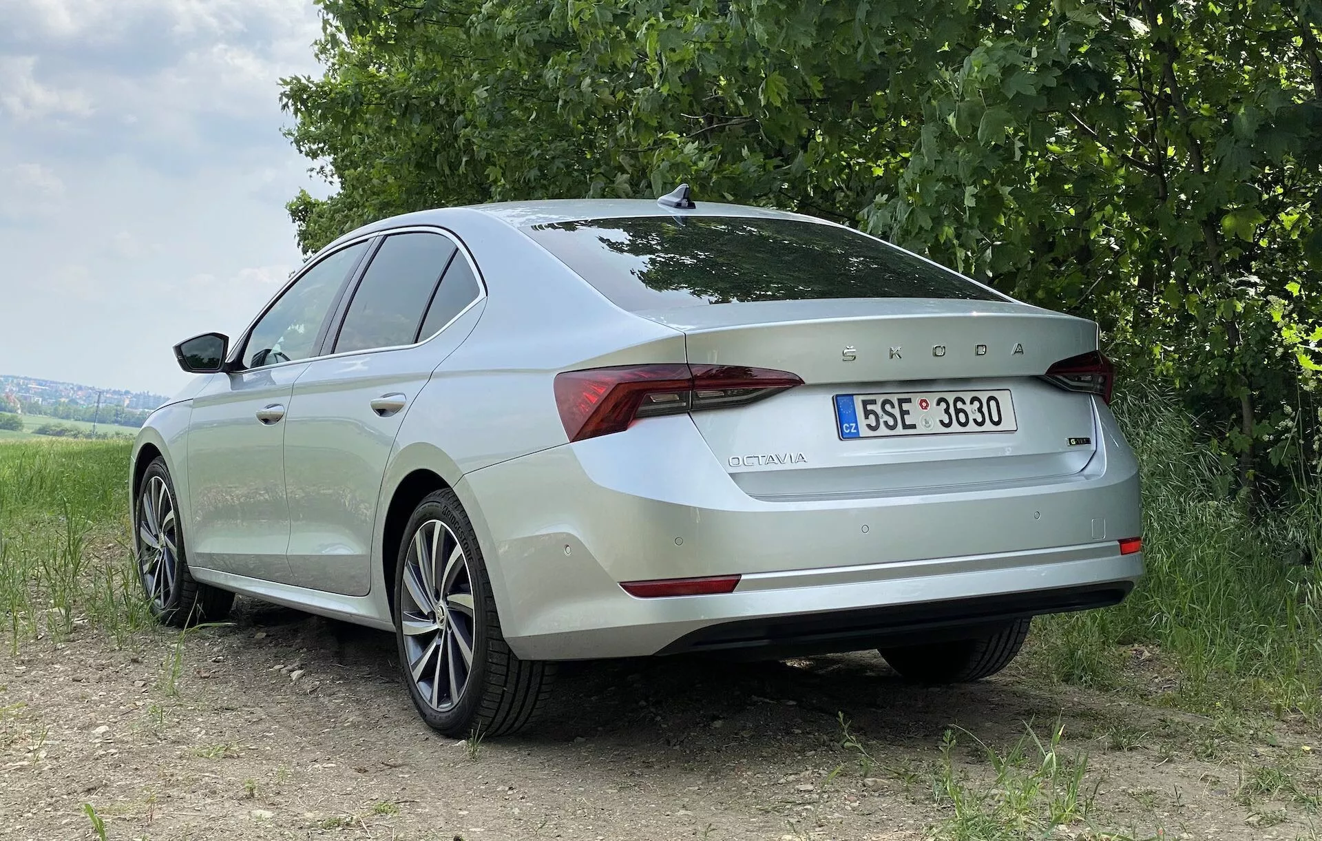 Vue trois-quarts arrière d'une berline Skoda Octavia 2020 de couleur claire sur une surface en terre, avec de l'herbe et des arbres en arrière-plan. La photo met en valeur le profil arrière et latéral du véhicule, montrant des lignes lisses, des surfaces galbées, des jantes en alliage et des feux arrière à LED élégants. L'emblème Skoda et le nom du modèle 'OCTAVIA' sont clairement visibles sur le couvercle du coffre, ainsi qu'une plaque d'immatriculation européenne. La lumière naturelle et douce accentue les éléments de design de la voiture sans ombres dures, suggérant un ciel nuageux ou une lumière solaire diffuse.
