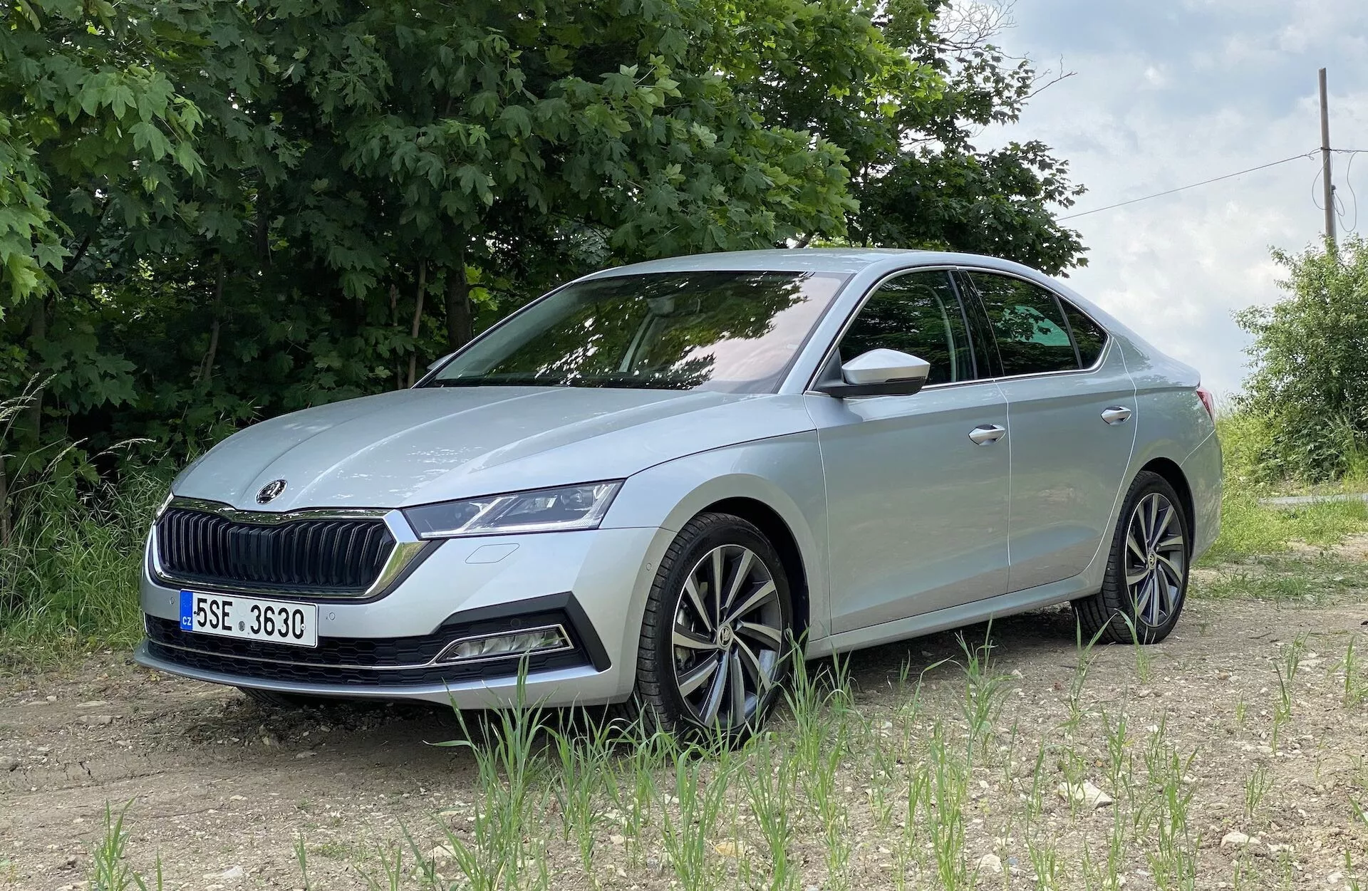 Skoda Octavia 2020 vue sous un angle de trois quarts avant bas, mettant en valeur le côté avant gauche détaillé incluant l'assemblage des phares à LED, la calandre proéminente, le pare-chocs avant avec capteurs, les lignes sculptées du capot, les roues en alliage à plusieurs rayons, le clignotant intégré dans le rétroviseur latéral et les vitres légèrement teintées, positionnée sur une surface non pavée avec du feuillage vert et une clôture en fil de fer dans un cadre rural ou semi-rural.