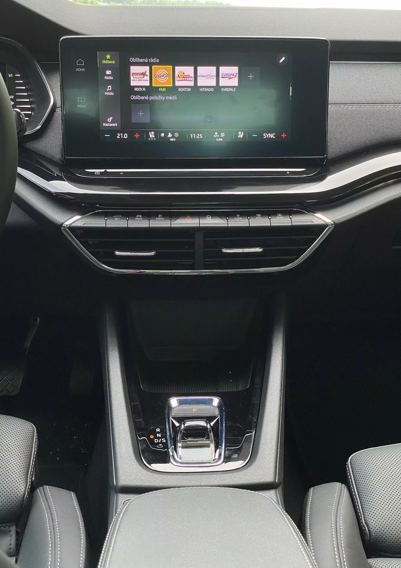 Perspective intérieure depuis l'aire des passagers arrière d'une Skoda Octavia limousine 2020, montrant le système d'infodivertissement à écran tactile avec des icônes colorées pour les médias, le téléphone et les paramètres du véhicule. En dessous se trouvent les boutons de contrôle du climat à double zone avec un affichage numérique de la température, encadrés par des bouches d'aération verticales et horizontales. La console centrale élégante présente un levier de vitesse noir brillant avec une poignée argentée, des compartiments de rangement avec tapis en caoutchouc, et une sellerie texturée visible sur les sièges avant sous une lumière naturelle.