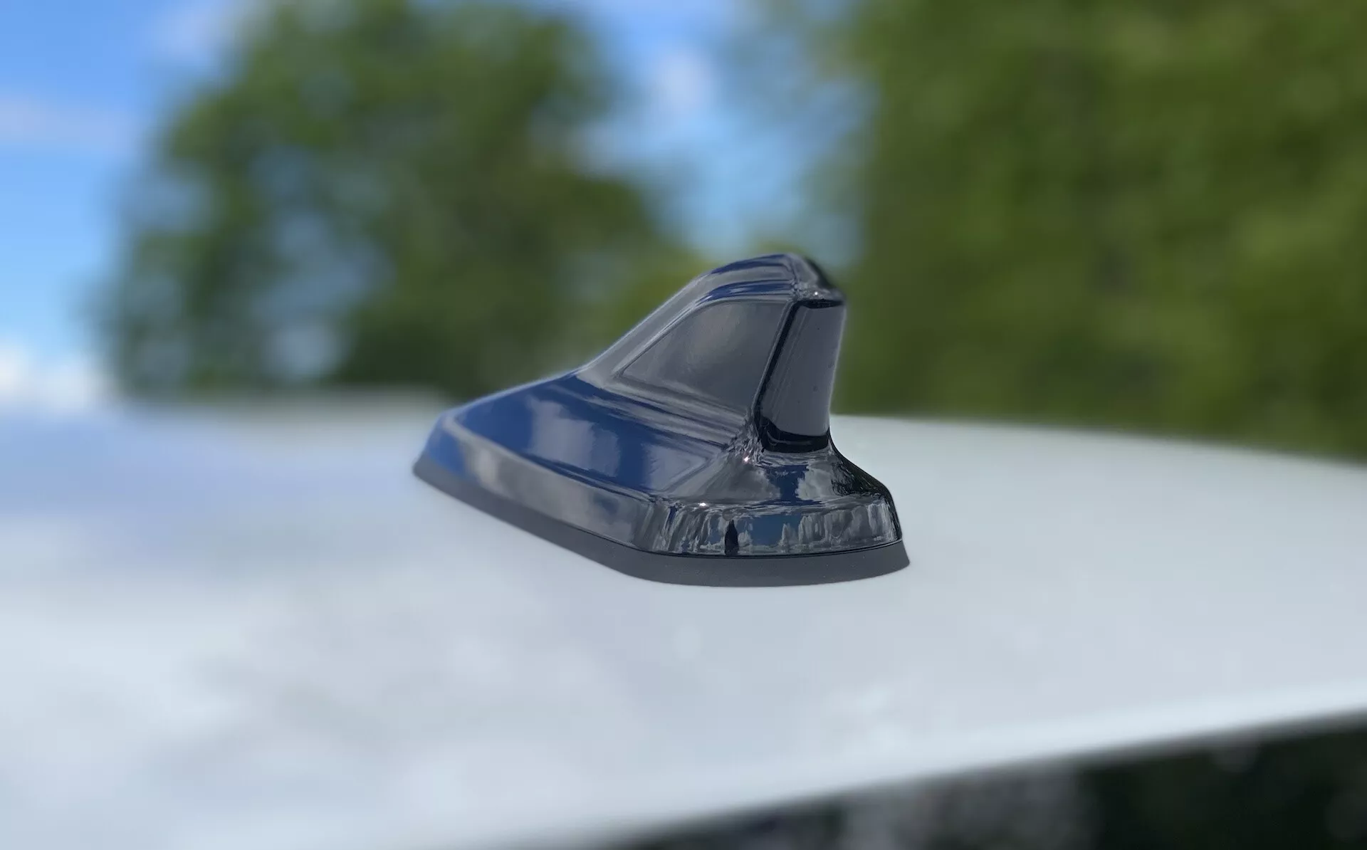 Gros plan sur une antenne de style aileron de requin sur le toit d'un véhicule, mettant en valeur un design automobile moderne. Prise de vue en contre-plongée avec appareil photo légèrement incliné vers le haut, mettant au point l'antenne aérodynamique et brillante sur un fond flou de feuillage et de ciel. Le design épuré suggère des fonctionnalités comme la radio FM/AM, la navigation par satellite et les technologies de communication avancées. La finition métallique reflète l'extérieur bien entretenu du véhicule.