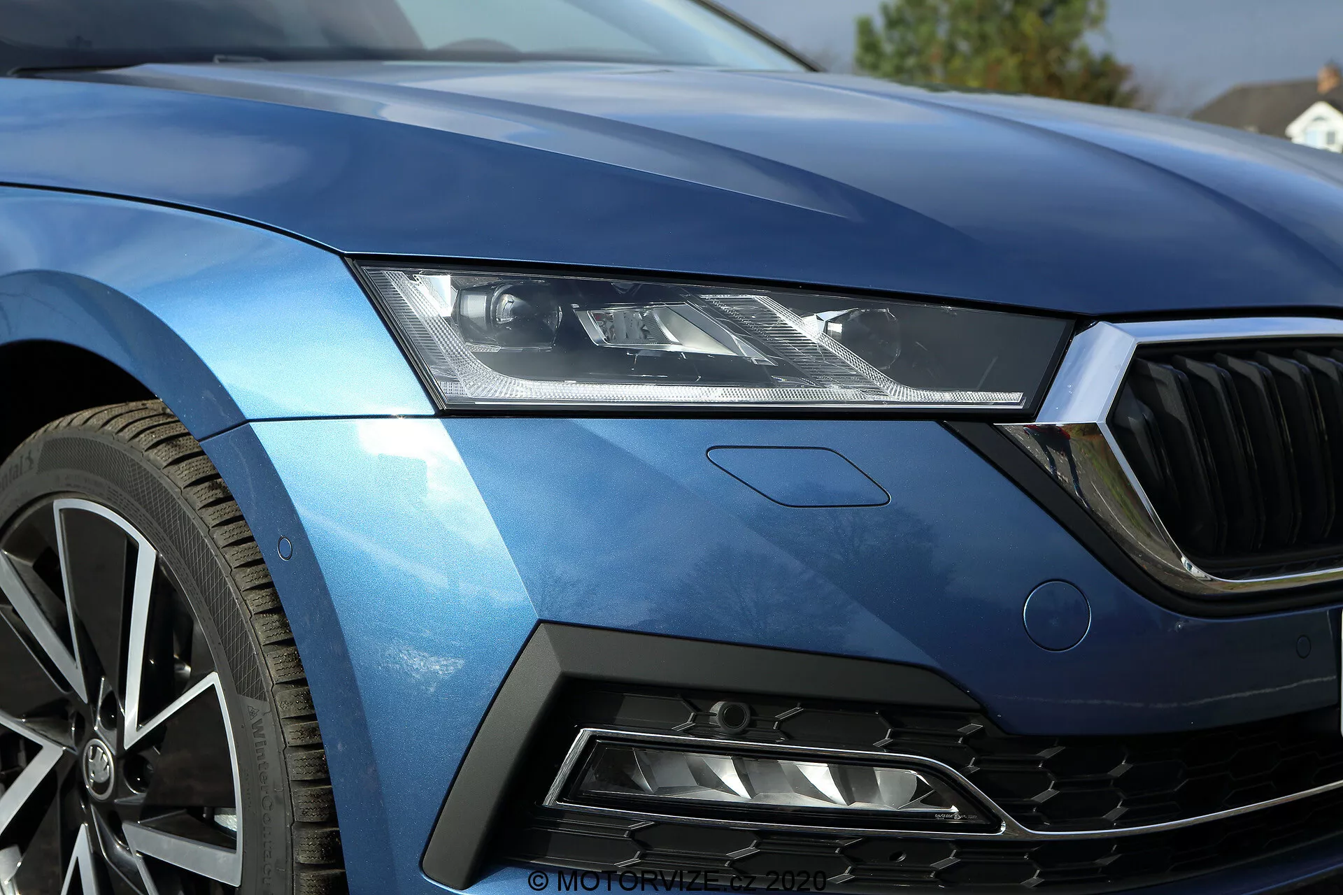 Gros plan du quart avant gauche d'une Skoda Octavia limousine 2020, montrant le groupe de phares à LED élégant, une partie du capot, le bord gauche de la calandre avant, l'aile avant gauche aérodynamique et un segment du pare-chocs avec une prise d'air à motif en nid d'abeille. La peinture bleue métallisée et les lignes nettes soulignent le luxe et le sport. La roue en alliage à plusieurs rayons suggère une haute performance. Le design comprend un badge subtil, indiquant un niveau de finition ou un paquet technologique spécifique. Cette image capture magnifiquement la technologie avancée des phares du véhicule, la finition de la peinture et les caractéristiques de conception axées sur la performance.