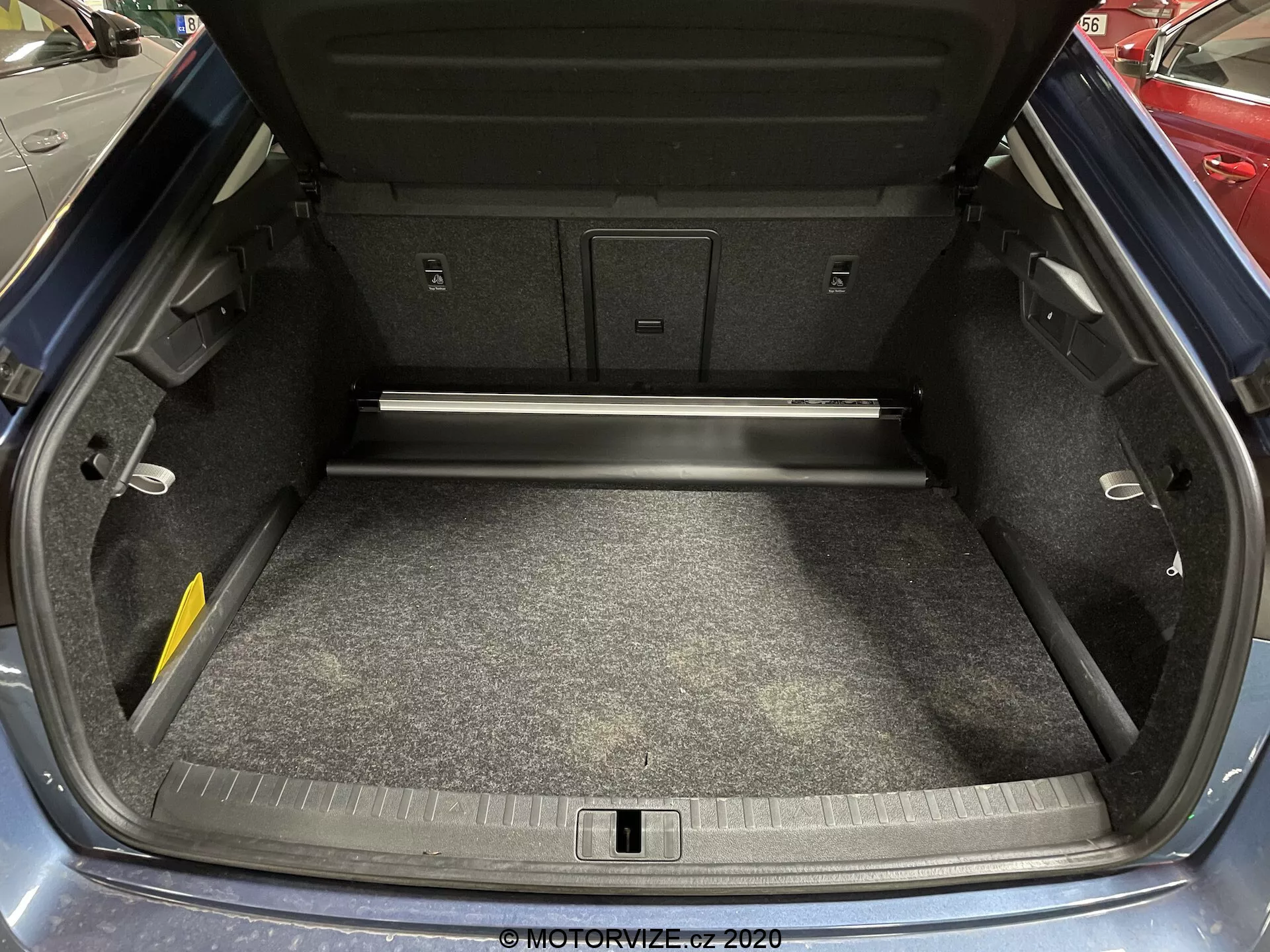 Image du coffre de la Skoda Octavia 2020 avec le couvercle ouvert, montrant un intérieur spacieux et propre, un sol en moquette, tapissé de panneaux en matériau composite, une fonctionnalité de passage arrière pour les objets longs et un crochet de chargement visible. Le pare-chocs arrière montre des marques d'usure mineures.