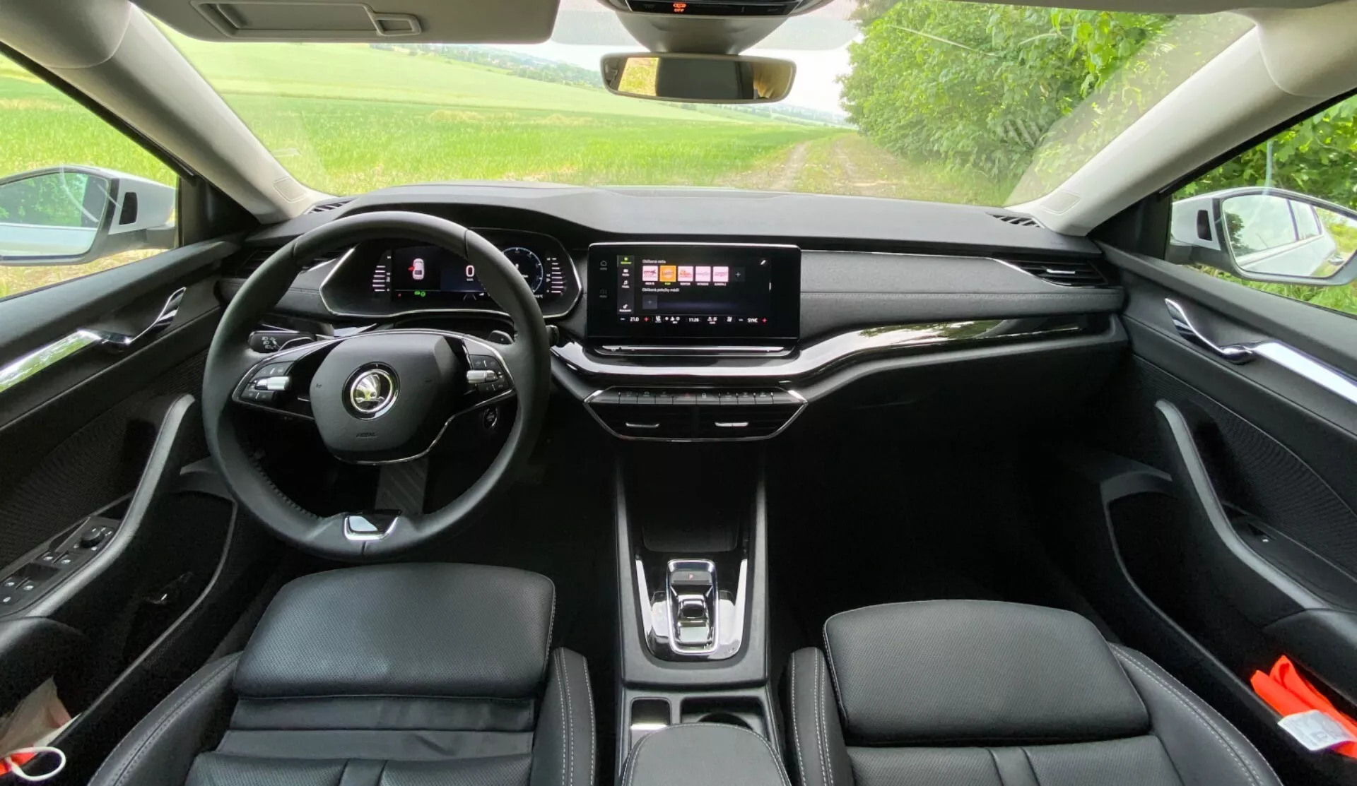 Vue intérieure d'une Skoda Octavia 2020 depuis la banquette arrière montrant le volant moderne à fond plat avec commandes intégrées, le tableau de bord numérique, un grand système d'infodivertissement à écran tactile sur un tableau de bord épuré, un minimum de boutons physiques, le pad de recharge sans fil, les sièges avant ergonomiques en similicuir avec surpiqûres contrastées et un toit ouvrant panoramique permettant à la lumière naturelle d'entrer dans l'habitacle.
