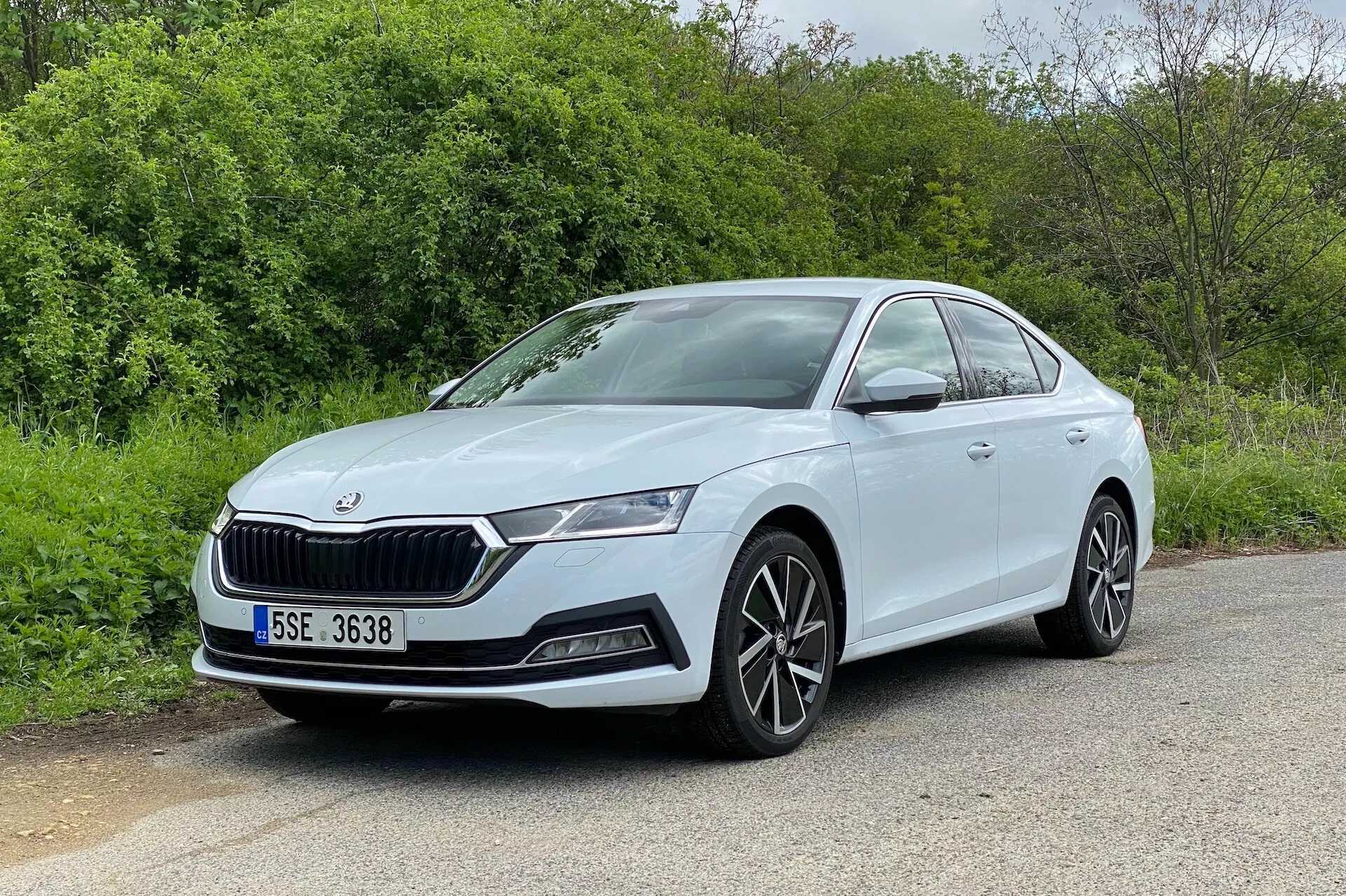 L'image montre une berline Skoda Octavia 2020 sous un angle de trois quarts avant, présentant à la fois la face avant et le profil latéral côté conducteur. La voiture, garée sur une surface asphaltée avec des éléments naturels comme des arbres ou des buissons en arrière-plan, possède une calandre distinctive, des phares à LED élégants, des logements pour phares antibrouillard et des prises d'air sur le pare-chocs. Les jantes en alliage multi-branches et les lignes latérales lisses et aérodynamiques rehaussent son apparence moderne et sportive. La vue bien éclairée et dégagée met en évidence les poignées de porte de la couleur de la carrosserie et la ligne de caractère visible sur les portes, en faisant une excellente représentation du design automobile.