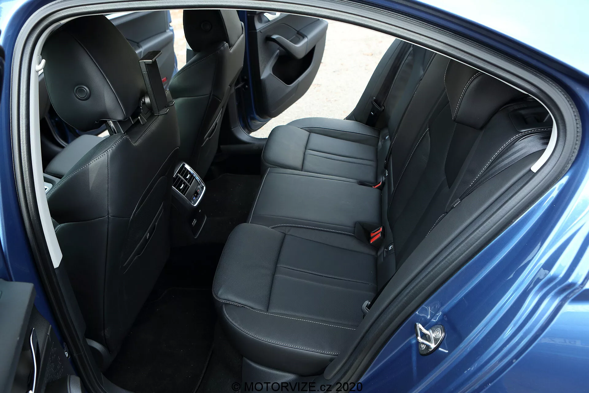 Vue aérienne du compartiment arrière des passagers d'une limousine Skoda Octavia 2020, prise du côté droit. La photo montre une porte ouverte avec des sièges modernes en cuir noir ou en similicuir avec des coutures de luxe. La banquette arrière est conçue pour accueillir confortablement deux passagers avec des appuie-têtes individuels, indiquant un modèle sportif ou orienté vers le confort. L'image met en évidence les bouches de ventilation de la climatisation sur la console centrale, les ports USB pour la recharge des appareils, les haut-parleurs du système audio avancé dans le panneau de la porte, les commandes des fenêtres électriques, les ceintures de sécurité visibles y compris une ceinture à trois points pour le siège central, et les tapis de sol en moquette. Le schéma de couleur sombre de l'intérieur indique un véhicule premium bien équipé sans objets personnels visibles, en position de stationnement avec un fond neutre.