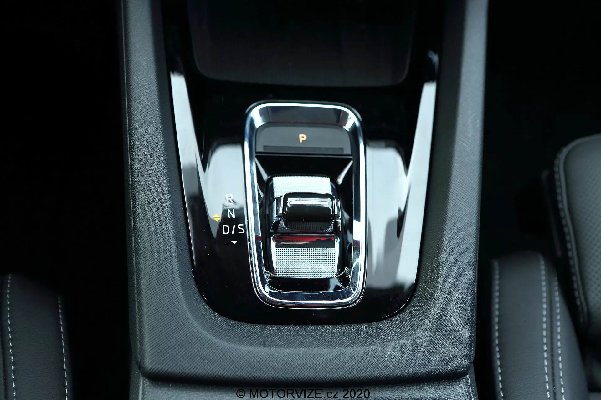 Vue rapprochée du levier de vitesses moderne dans la console centrale de la Skoda Octavia Limousine 2020. Le levier de vitesses de face présente un design élégant et rectangulaire avec des bords arrondis et un contour chromé, affichant les positions de vitesses 'P', 'N', 'D/S'. Entouré de matériaux noirs de haute qualité avec des coutures visibles et une surface argentée avec une texture en maille fine, suggérant luxe et détail. Un éclairage lumineux illumine l'intérieur robuste et cohésif, indicatif de fonctionnalités électroniques avancées et de technologies automobiles modernes.