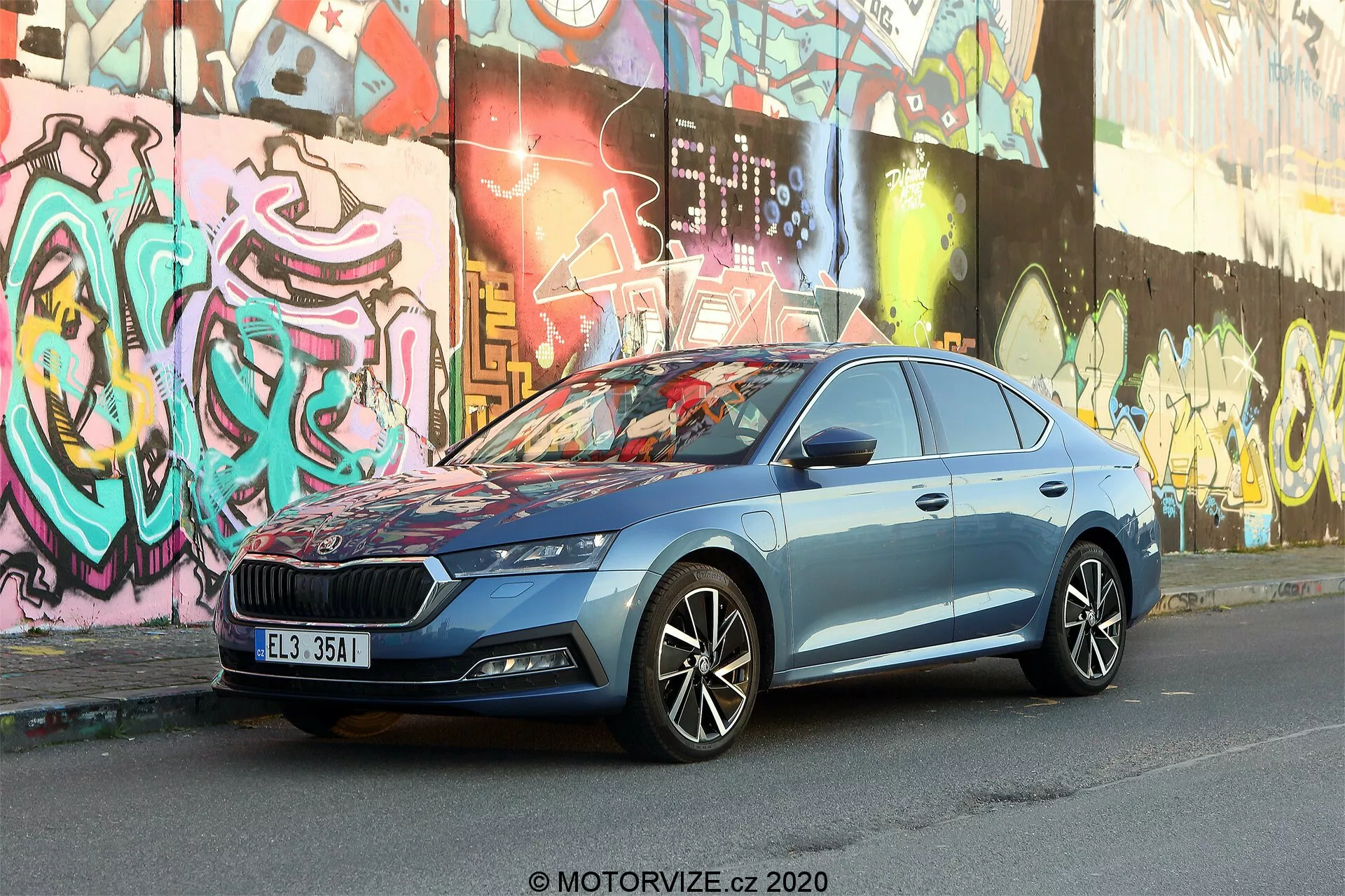L'image présente une Skoda Octavia Limousine 2020 garée dans une rue urbaine avec des murs couverts de graffitis en arrière-plan. Capturée sous un angle trois-quarts avant légèrement bas, elle met en avant la calandre substantielle de la voiture avec un motif vertical et un emblème central, des phares LED élégants, des jantes en alliage multi-branches, des lignes de carrosserie dynamiques, des rétroviseurs latéraux avec clignotants intégrés, des fenêtres encadrement chromé et une peinture bleue métallisée. L'environnement urbain souligne le design moderne et la technologie automobile avancée du véhicule.