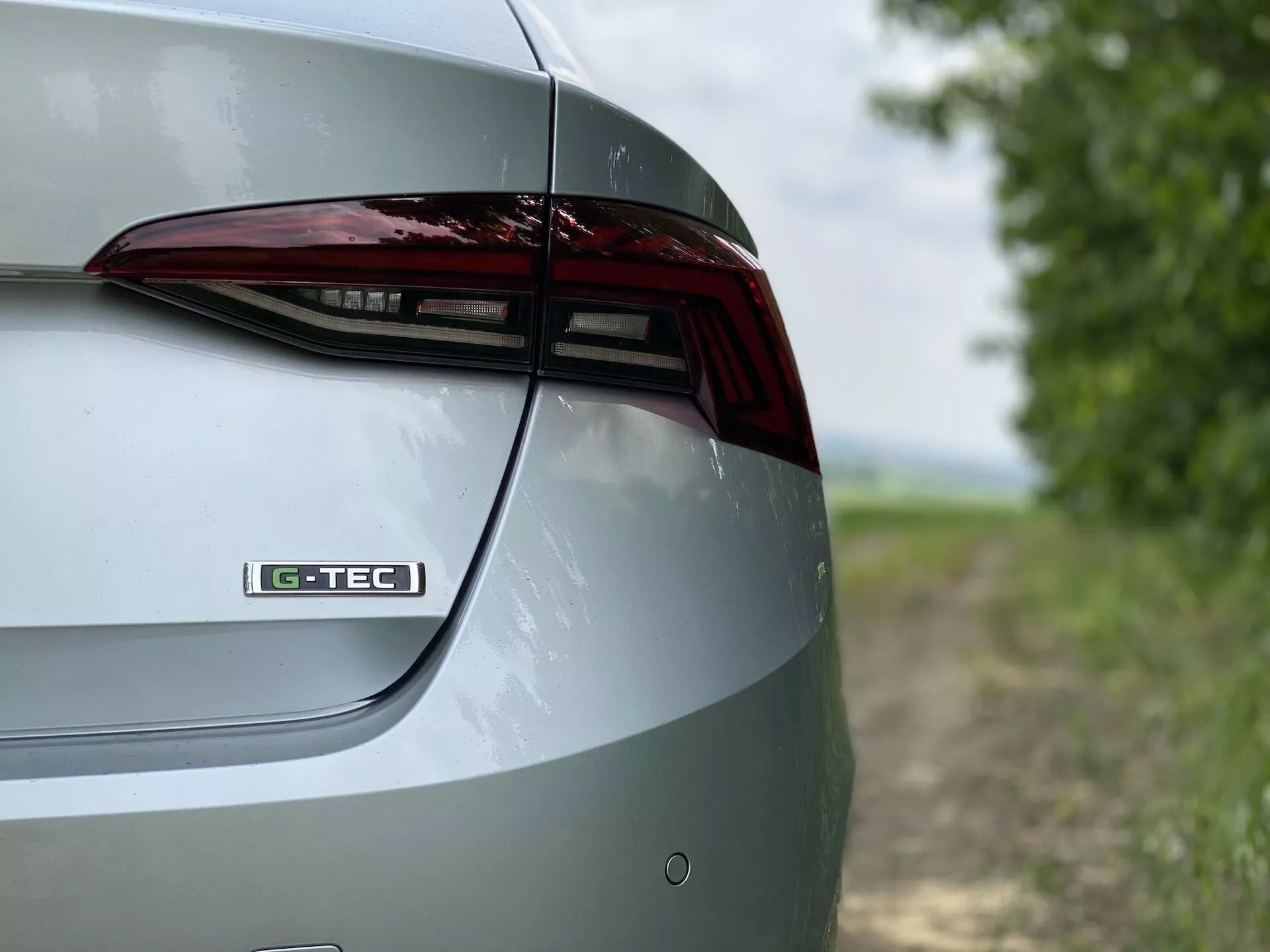 Vue détaillée en gros plan de la section arrière droite d'une Skoda Octavia 2020 berline. La prise de vue capture les éléments de design du couvercle du coffre, du feu arrière à LED et d'une partie du pare-chocs arrière sous un angle oblique et légèrement surélevé. Le badge 'G-TEC' sur le couvercle du coffre suggère une technologie ou une finition spécifique. La finition métallique de la peinture met en valeur l'extérieur poli de la voiture. L'arrière-plan flou crée un cadre rural qui met en avant les caractéristiques et le design du véhicule.