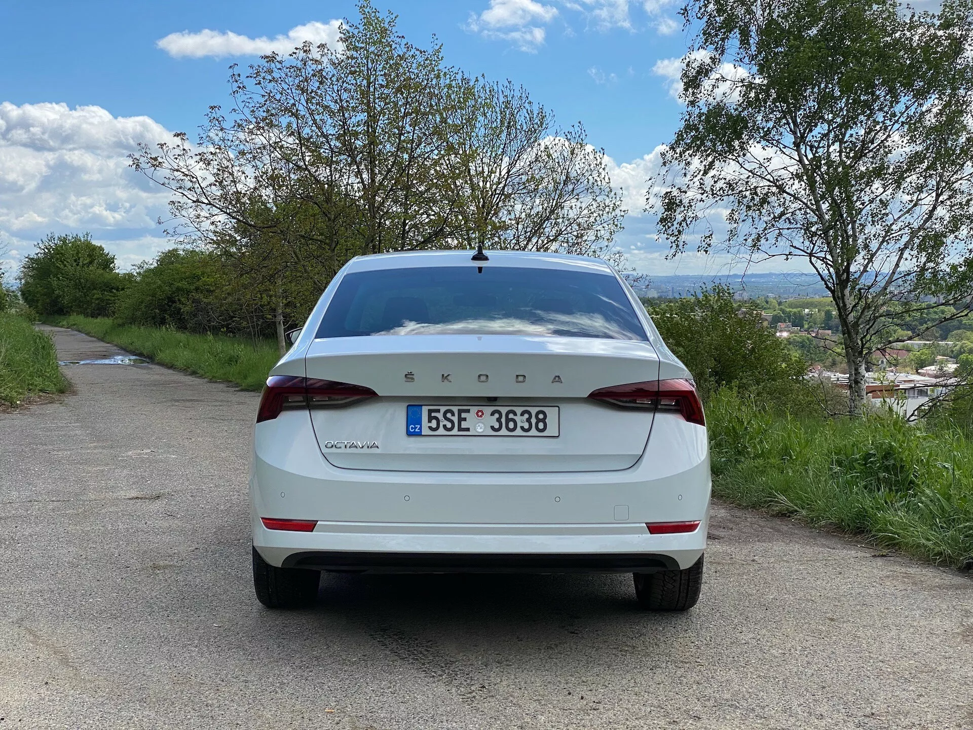 Vue arrière d'une Škoda Octavia 2020 berline blanche, stationnée sur une route asphaltée étroite dans un cadre rural. L'image, prise au niveau du sol avec un angle droit direct, montre le couvercle du coffre, le pare-chocs arrière, les feux arrière, la plaque d'immatriculation, le pare-brise arrière et la marque Škoda répartie au centre du couvercle de coffre avec 'Octavia' sur la gauche. Autour du véhicule, on voit des arbres et un ciel bleu nuageux, avec un village ou un paysage lointain visible sous l'horizon.