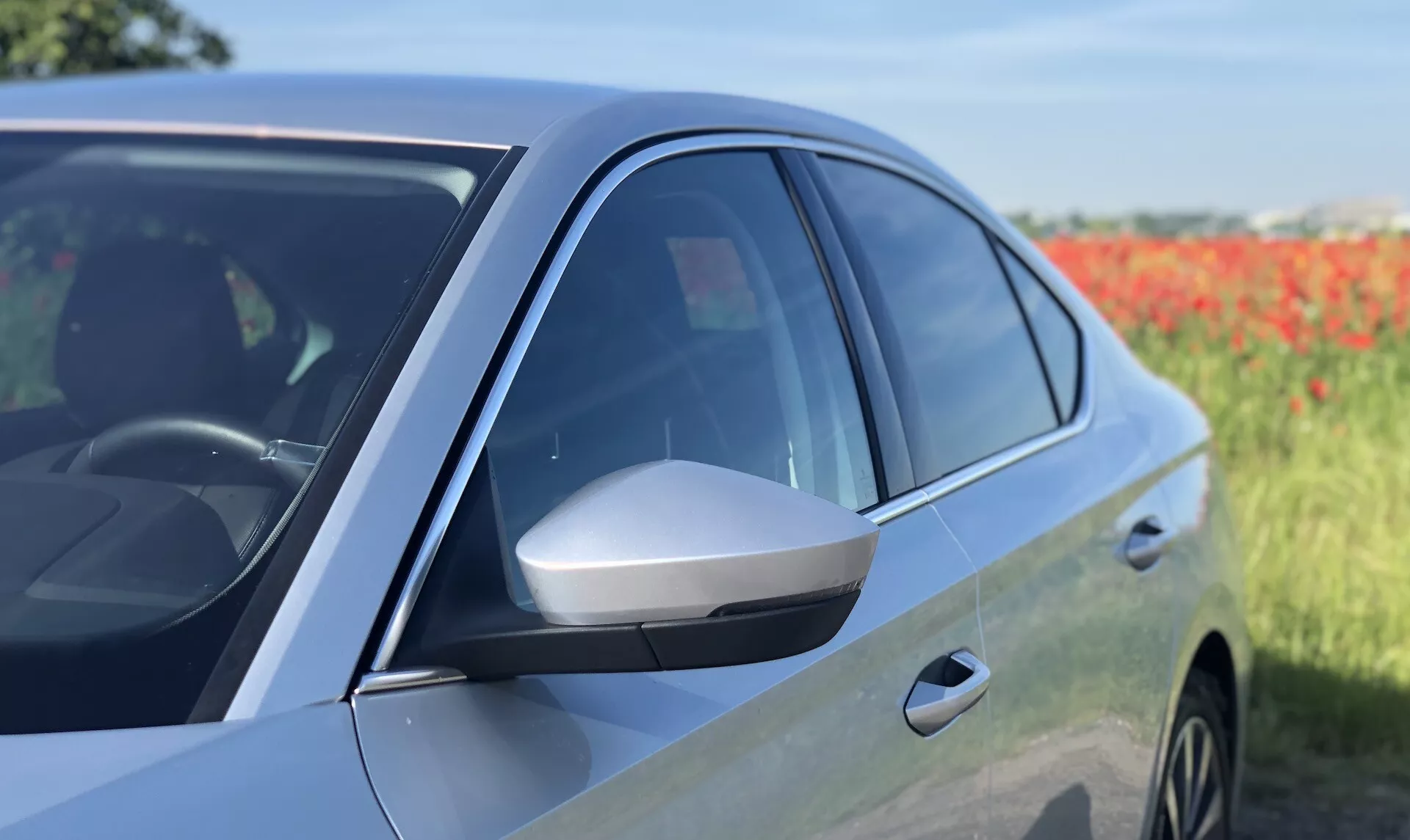 Vue latérale en gros plan d'une berline moderne Škoda Octavia 2020, montrant la porte avant côté conducteur et le rétroviseur latéral. L'image met en avant le rétroviseur latéral profilé avec clignotant intégré, les vitres teintées, la finition de peinture brillante et le design aérodynamique du milieu vers l'avant. Le reflet dans le panneau de la porte et une partie de la roue avant suggèrent des jantes en alliage. La photo met en évidence des caractéristiques de design avancées et les technologies potentielles du véhicule sur un fond naturel légèrement flou.