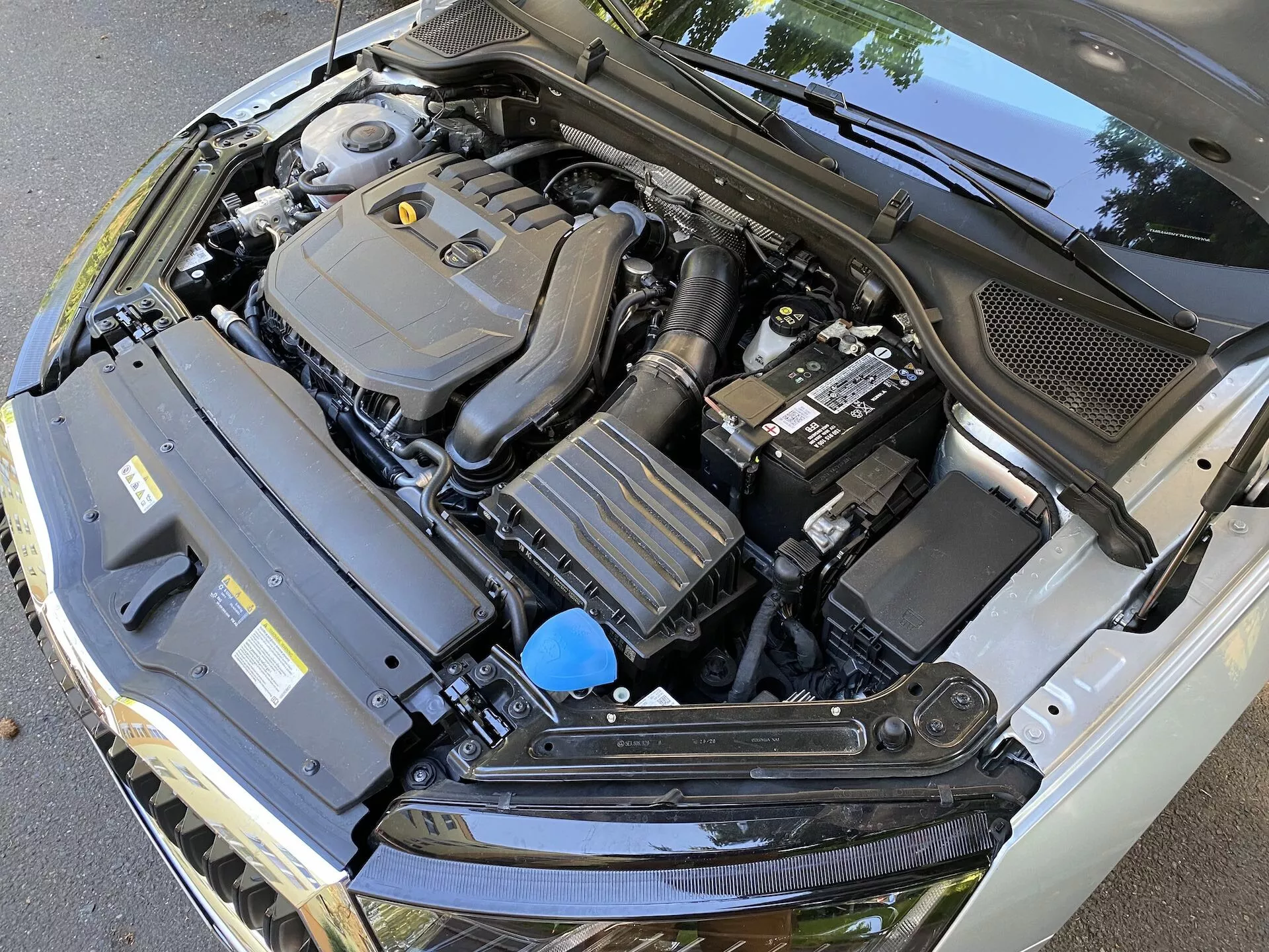 Vue en gros plan du compartiment moteur de la Skoda Octavia 2020 limousine avec le capot ouvert, montrant divers composants, y compris le couvercle du moteur encadré au centre, la batterie à droite, un composant rectangulaire avec ailettes de refroidissement à gauche et un réservoir de liquide avec un bouchon bleu. L'image, prise sous un angle descendant légèrement décentré, montre également le panneau en plastique noir à la base du pare-brise, soulignant la complexité technique du compartiment moteur automobile.