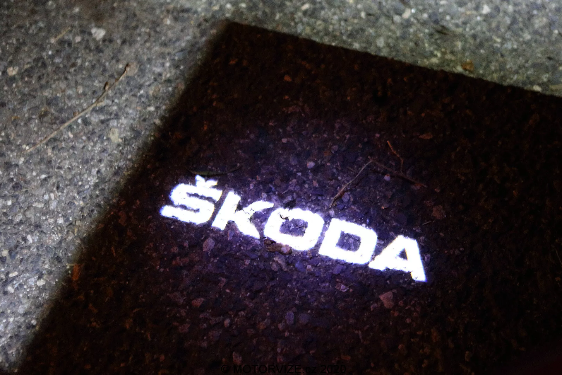 Projection du logo Škoda sur le sol. La source lumineuse, probablement située à l'extérieur de la Škoda Octavia 2020 (octavia-limousine), crée une 'lumière de flaque' qui éclaire le sol lorsque la porte du véhicule est ouverte. Cette projection de lumière blanche nette et claire, sur fond sombre, sert à la fois des fins fonctionnelles et de branding, indiquant la présence de technologie LED moderne dans les accessoires du véhicule.