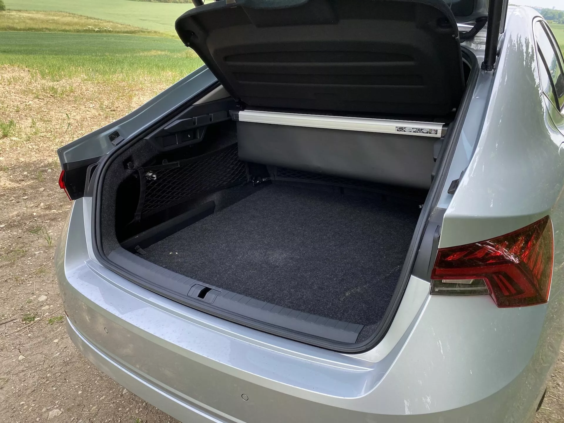 Vue en gros plan à trois quarts du compartiment du coffre arrière d'une Škoda Octavia 2020, mettant l'accent sur l'espace de rangement ouvert avec un intérieur propre et moquetté. L'angle capture une partie du côté droit et arrière de la voiture, montrant des feux arrière de conception moderne, une peinture métallisée lisse et un pare-chocs partiellement visible. La partie inférieure du couvercle du coffre comprend une poignée ergonomique, et une étagère est visible en haut de l'espace de chargement. L'environnement suggère un cadre extérieur avec un éclairage doux en journée.