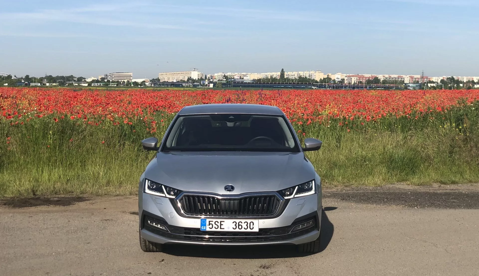 Vue avant d'une Skoda Octavia Limousine 2020 sur une route ou une piste de terre, montrant sa façade avant avec une calandre distinctive en forme de rein flanquée de phares LED ou adaptatifs élégants. Le pare-chocs avant aérodynamique présente des découpes pour phares antibrouillard et des prises d'air. Le capot présente des lignes de contour pour un design esthétique et aérodynamique. Le pare-brise entièrement visible peut suggérer des caractéristiques technologiques avancées comme les affichages tête haute ou les systèmes d'assistance à la conduite. Une voiture de couleur claire avec une plaque d'immatriculation montée au centre et des capteurs de stationnement, sur un fond de fleurs rouges.