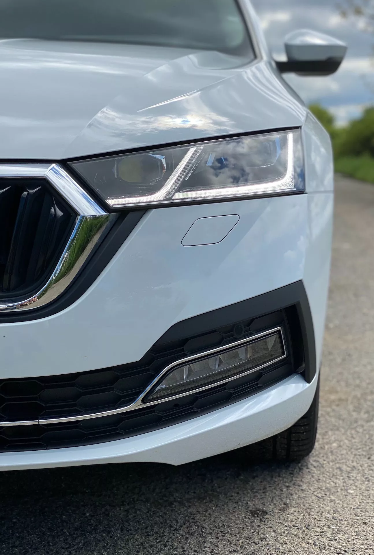 Vue rapprochée du quart avant gauche d'une Skoda Octavia 2020 (octavia-limousine), mettant en avant le phare à LED, la calandre distinctive avec des détails chromés, le pare-chocs avant moderne avec prise d'air et capteurs, le design du capot avec un pli et le rétroviseur latéral partiellement visible avec clignotants intégrés. La voiture est peinte dans une couleur claire, probablement blanche ou argentée, avec des lignes de carrosserie élancées. L'image est prise d'une perspective basse, se concentrant sur le phare et la calandre, avec une faible profondeur de champ qui floute les éléments environnants.