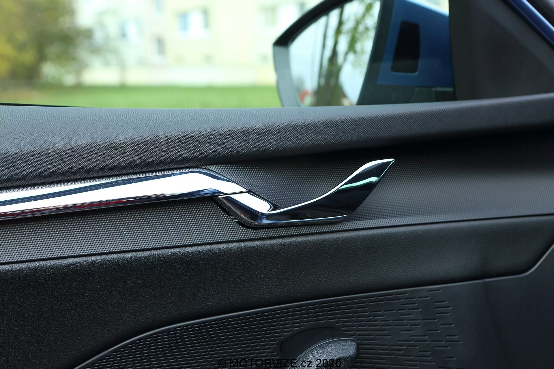 Vue intérieure en gros plan d'une Skoda Octavia 2020 se concentrant sur l'arrière de la porte avant du passager, mettant en évidence la poignée de porte chromée réfléchissante. Le panneau de porte présente un matériau en similicuir ou à texture souple avec des coutures visibles, indiquant une finition intérieure haut de gamme. Également visible est une grille de haut-parleur faisant partie du système audio de la voiture, avec un motif uniforme et un design coordonné en termes de couleurs. L'image est prise depuis le siège arrière, suggérant que la voiture est garée à l'extérieur dans une zone résidentielle, avec la verdure floue en arrière-plan.