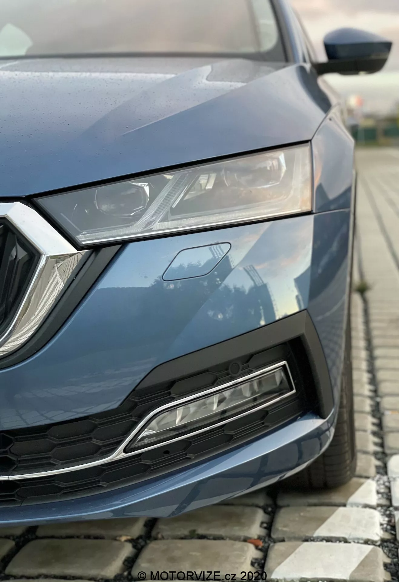 Image partielle en gros plan d'une Skoda Octavia 2020 moderne capturée sous un angle bas, se concentrant sur la section avant-gauche incluant capot, phare LED élégant, grille géométrique et pare-chocs avant avec prise d'air. Peinture bleue métallisée, éléments de design angulaires et caractéristiques aérodynamiques mises en avant.