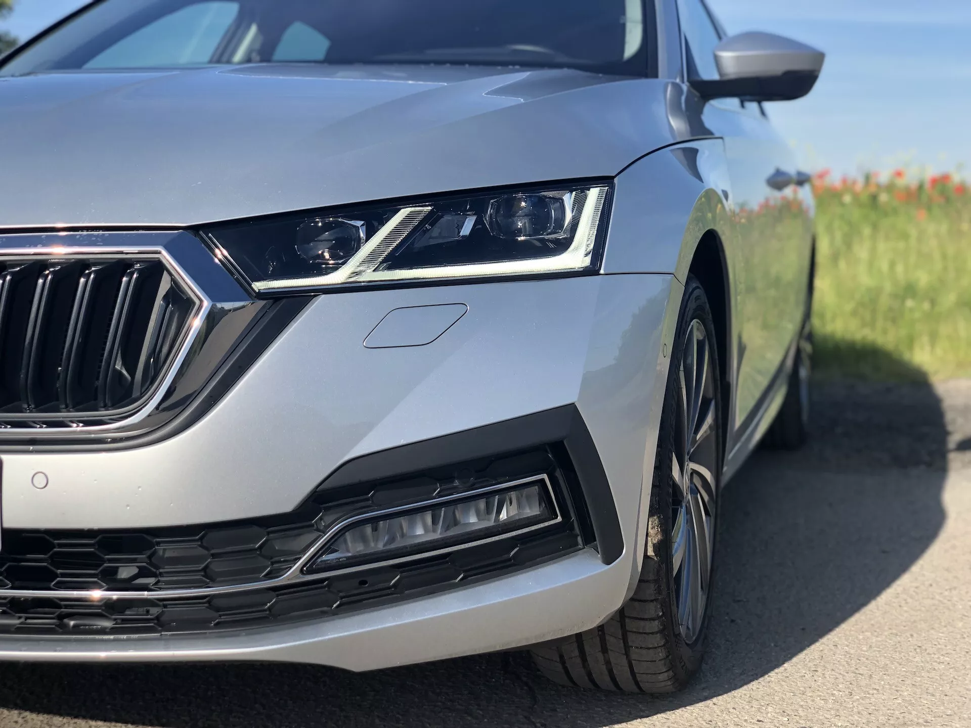 Vue rapprochée du quart avant gauche d'une limousine Skoda Octavia argentée de 2020, mettant en valeur le phare LED élégant, la calandre finition chrome avec des lamelles verticales, l'entrée d'air sculptée ainsi que la roue et l'aile avant partiellement visibles. La prise de vue est réalisée sous un angle bas au niveau du sol, se concentrant sur les éléments de design moderne et les caractéristiques technologiques avancées avec un arrière-plan flou.