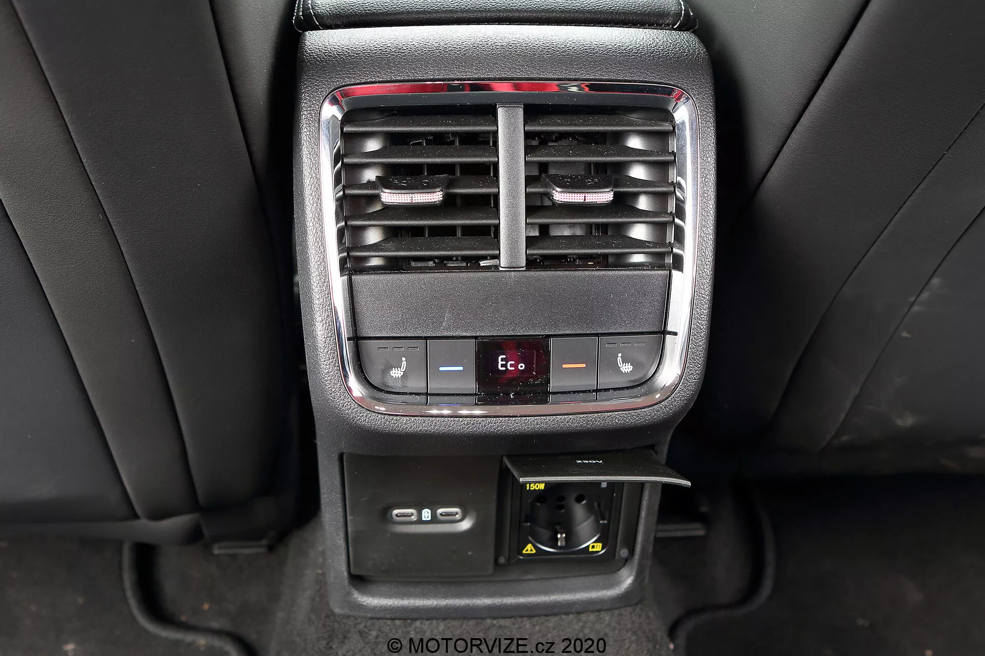 L'image montre la console centrale intérieure d'une Skoda Octavia Limousine 2020, mettant en évidence les caractéristiques de contrôle du climat et de commodité pour les passagers arrière. La photo offre une vue claire des aérateurs réglables, des boutons de contrôle de la température, d'un bouton mode Éco, d'une prise de courant 12V et d'un vide-poche de rangement.