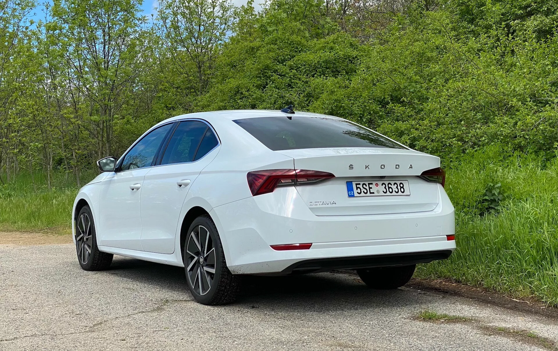 L'image montre une berline Škoda Octavia 2020 blanche prise sous un angle arrière trois quarts, mettant en évidence les composants du côté et de l'arrière droit du véhicule. Les détails clés incluent les feux arrière à LED, le logo Škoda et le badge du modèle OCTAVIA, la désignation TDI, le couvercle de coffre sculpté avec spoiler intégré, les jantes en alliage à cinq branches, la ligne de toit lisse en pente, le pare-chocs arrière avec capteurs de stationnement et la peinture brillante et bien entretenue. La voiture est garée sur une route rurale ou de parc tranquille avec de la verdure des deux côtés, renforçant le cadre serein. Cette photo met en avant le design extérieur de la voiture, les caractéristiques modernes de la berline et les éléments technologiques tels que le moteur diesel et les systèmes d'assistance au conducteur.