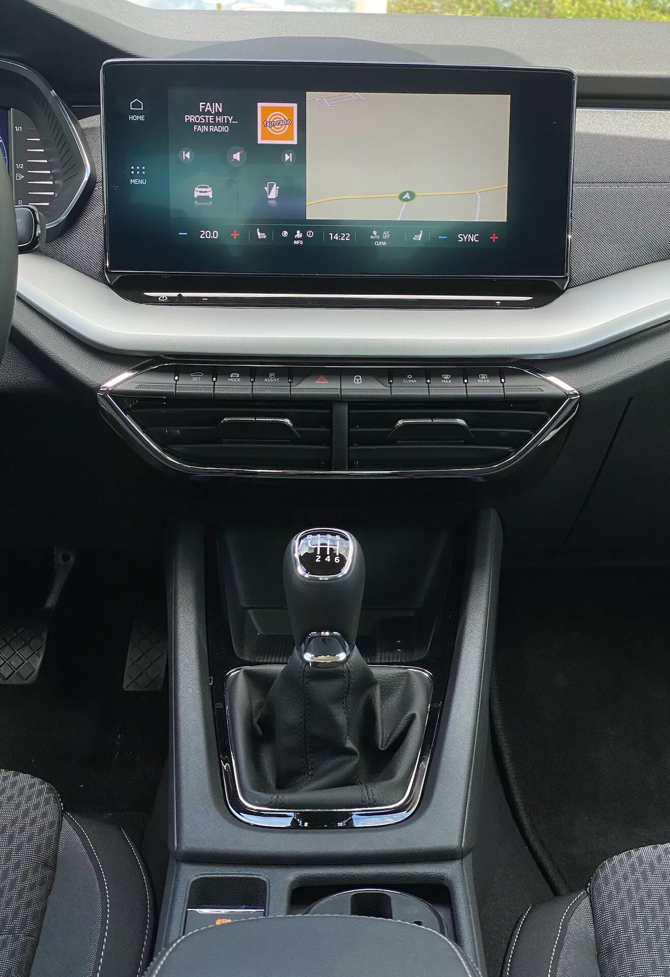 Vue détaillée de la console centrale intérieure d'une Skoda Octavia limousine 2020 depuis la perspective du siège arrière, montrant un grand écran tactile d'infotainment avec des commandes de médias et de climatisation, des bouches d'aération avec des accents métalliques et des boutons de conception minimaliste, un levier de vitesses automatique de style manuel traditionnel avec des signes de mode manuel, un compartiment de rangement avec des ports USB, des porte-gobelets et une finition en cuir et plastique lisse mettant l'accent sur le confort moderne et la convivialité centrée sur le conducteur.