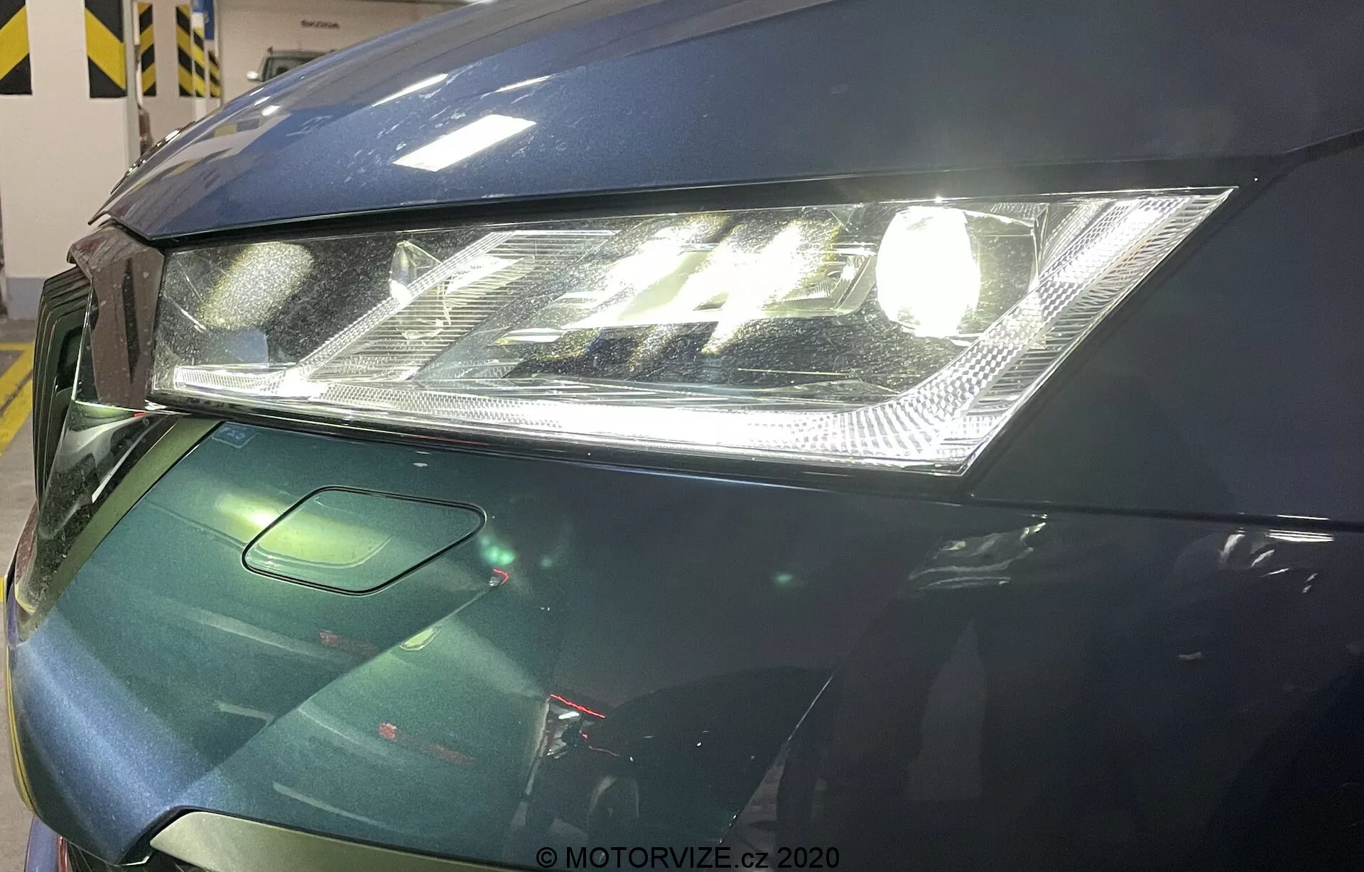 Vue rapprochée de la zone avant gauche d'une Skoda Octavia 2020. L'image se concentre sur un phare moderne avec lentille claire et feux de jour LED intégrés, un capot partiellement visible avec un design lisse, et une petite section de la calandre intégrant probablement des capteurs d'assistance au conducteur. La voiture présente une finition brillante de couleur vert foncé, suggérant des mesures avancées de protection de la peinture.