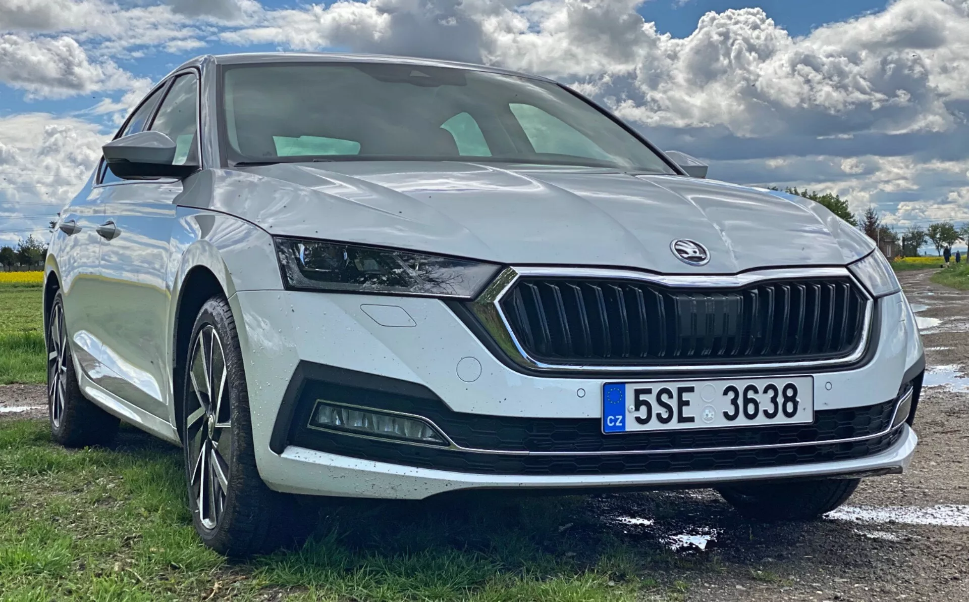 Vue en contre-plongée de trois quarts avant d'une Škoda Octavia 2020 blanche garée à l'extérieur, mettant en avant la face avant, le design angulaire des phares avec feux de jour à LED, la large calandre avec logo Škoda, le capteur de stationnement, le pare-chocs avant sportif avec phares antibrouillard, les jantes en alliage à plusieurs branches, et le clignotant intégré dans le rétroviseur latéral. Le pare-brise panoramique et la plaque d'immatriculation de style européen sont également visibles.