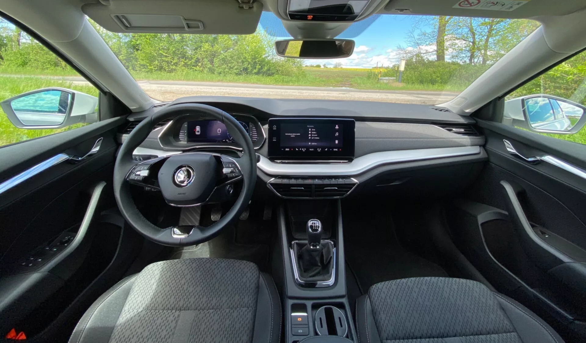 Vue intérieure de la Skoda Octavia 2020 depuis la perspective du conducteur, présentant le volant multifonction, le tableau de bord numérique, l'écran tactile d'infodivertissement horizontal, la console centrale minimaliste avec sélecteur de vitesse, porte-gobelets et tableau de bord tactile doux avec garniture imitant le bois sous la lumière directe du jour.