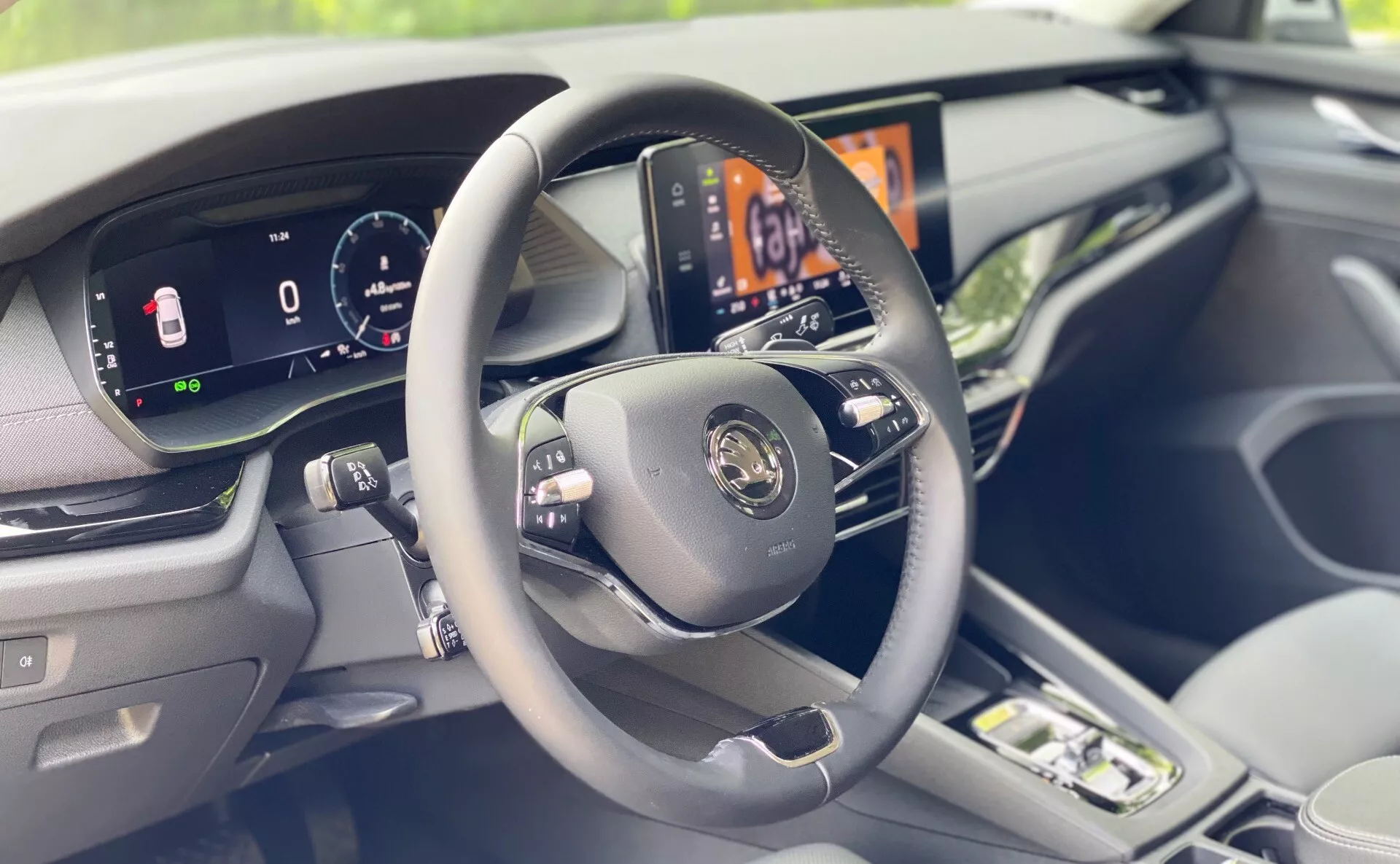 Vue intérieure d'un véhicule moderne focalisée sur le tableau de bord côté conducteur depuis une perspective légèrement à droite du siège du conducteur. L'image présente en premier plan un volant à trois branches avec des boutons de commande, un tableau de bord entièrement numérique affichant la vitesse du véhicule et d'autres informations essentielles, et un écran d'infodivertissement central haute résolution affichant le système de contrôle de la climatisation de la voiture. Les matériaux de haute qualité et les garnitures visibles sur le tableau de bord et la porte renforcent le sentiment de luxe de l'intérieur de la voiture. Le premier plan flou suggère la présence d'un siège passager.