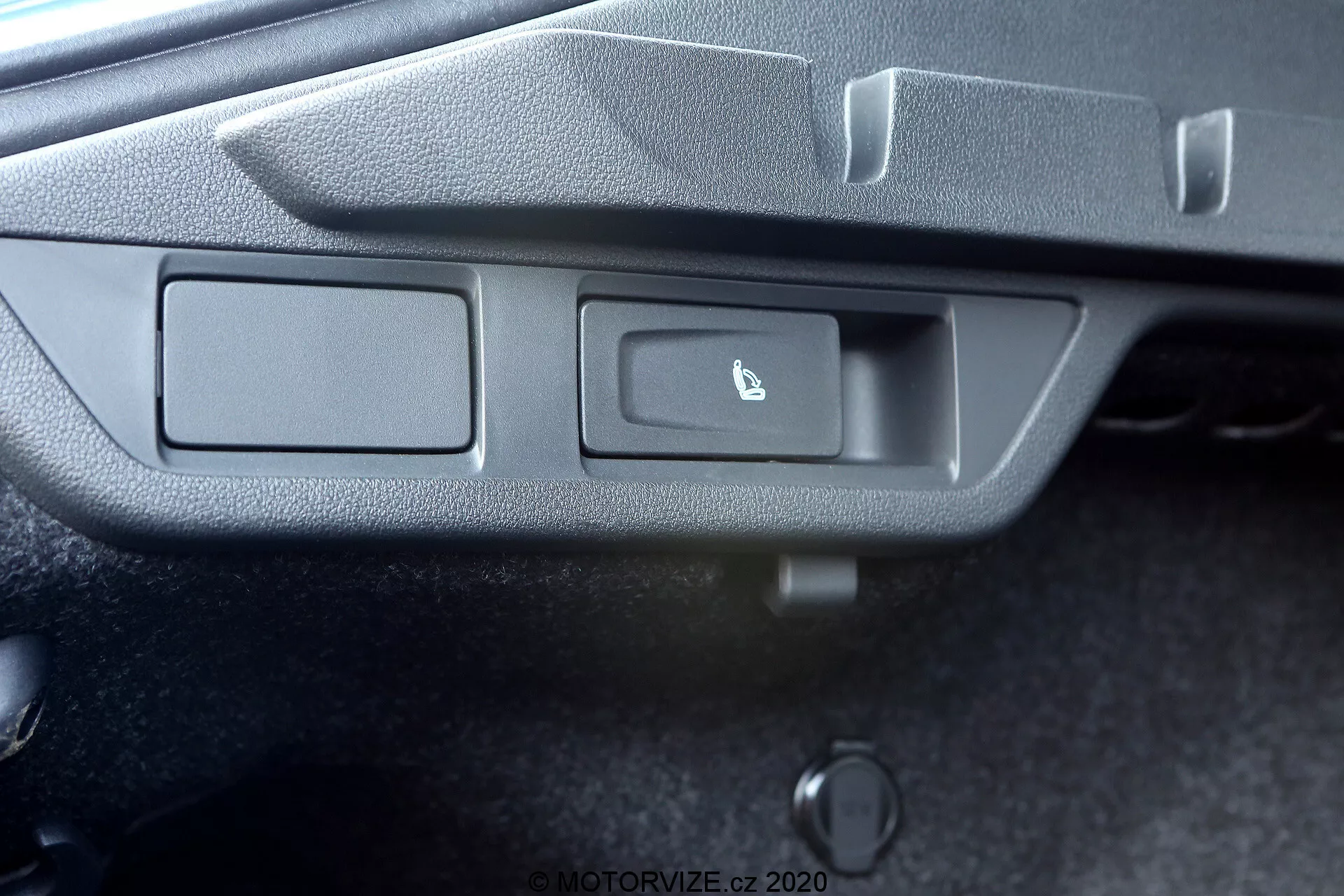 L'image capture une vue détaillée du panneau de commande d'une voiture sous un angle légèrement incliné. Le panneau comporte deux boutons rectangulaires encastrés dans un cadre en plastique gris texturé. Le bouton à gauche n'a pas de marque, tandis que celui de droite arbore une icône de coffre entre-ouvert, indiquant une fonction de déverrouillage du coffre. Une petite lampe témoin, probablement une LED, est située à droite des boutons. Au-dessous des boutons se trouve un objet cylindrique, possiblement un déverrouillage manuel ou d'urgence du coffre. La photo ne révèle pas le contexte plus large de l'intérieur de la voiture.