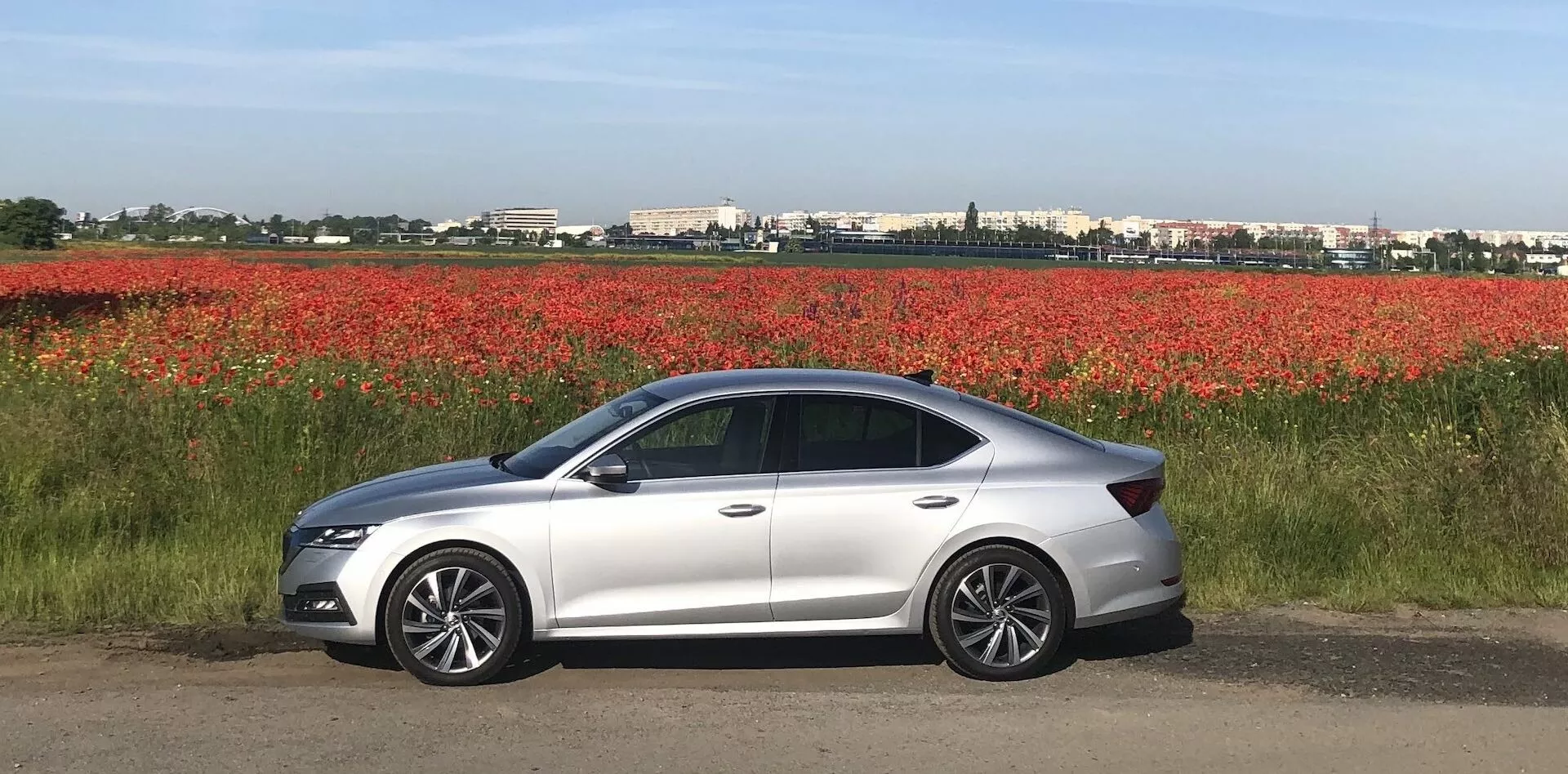 Vue latérale d'une berline Skoda Octavia 2020 moderne argentée garée sur une surface plane à côté d'un champ de fleurs rouges, présentant un design aérodynamique élégant, des phares à LED, des rétroviseurs couleurs carrosserie avec clignotants intégrés, des roues en alliage, quatre portes, des vitres teintées et une ligne de toit doucement inclinée.
