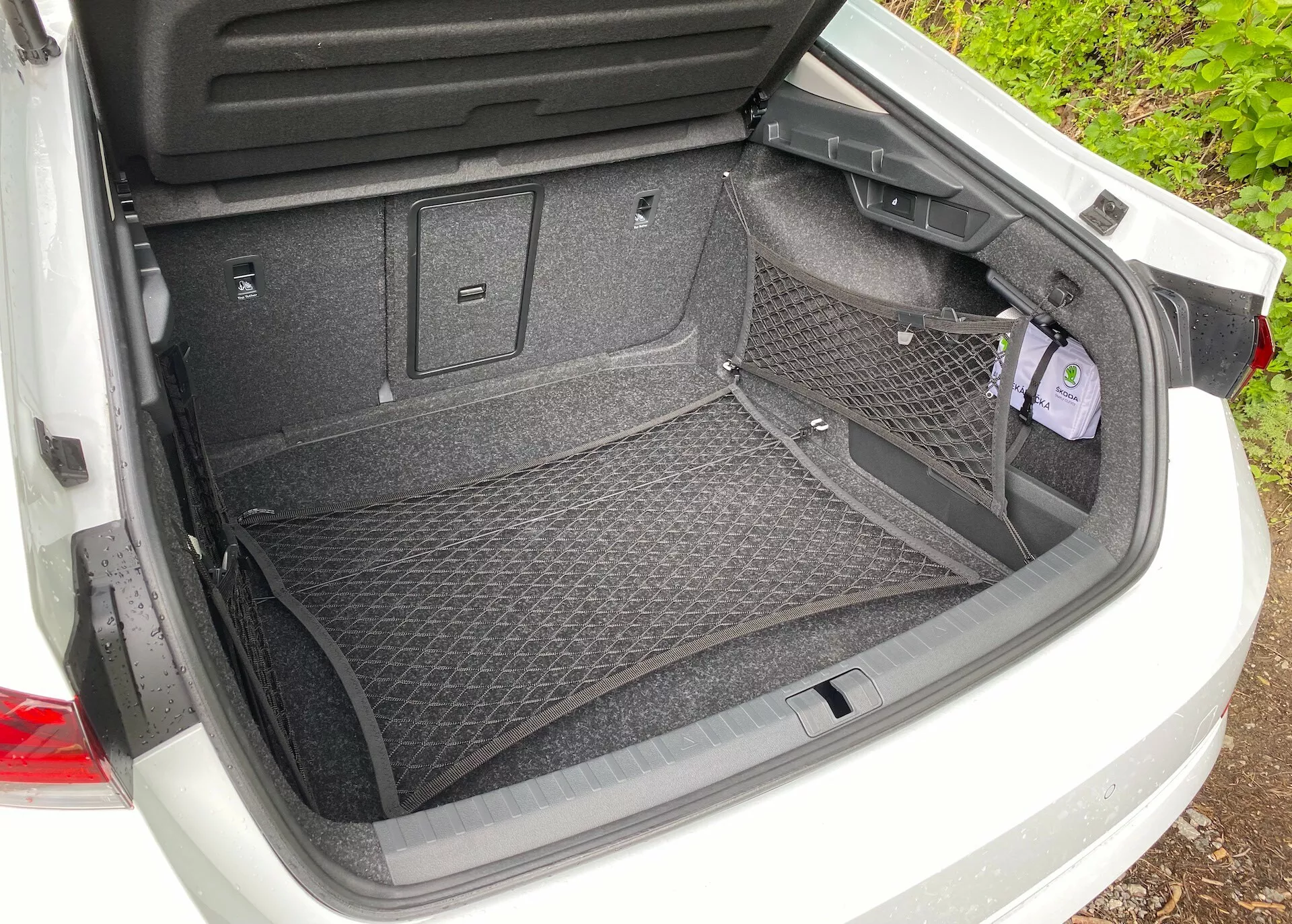 Vue arrière du coffre ouvert d'une Škoda Octavia 2020 (octavia-limousine) avec une surface en tapis et un accessoire filet de chargement pour un rangement sécurisé. Les caractéristiques comprennent un revêtement en plastique pour la protection, divers crochets et points d'attache pour des options de chargement supplémentaires. Un petit compartiment encastré à droite avec un couvercle indique un espace de rangement supplémentaire ou l'accès à des outils essentiels.