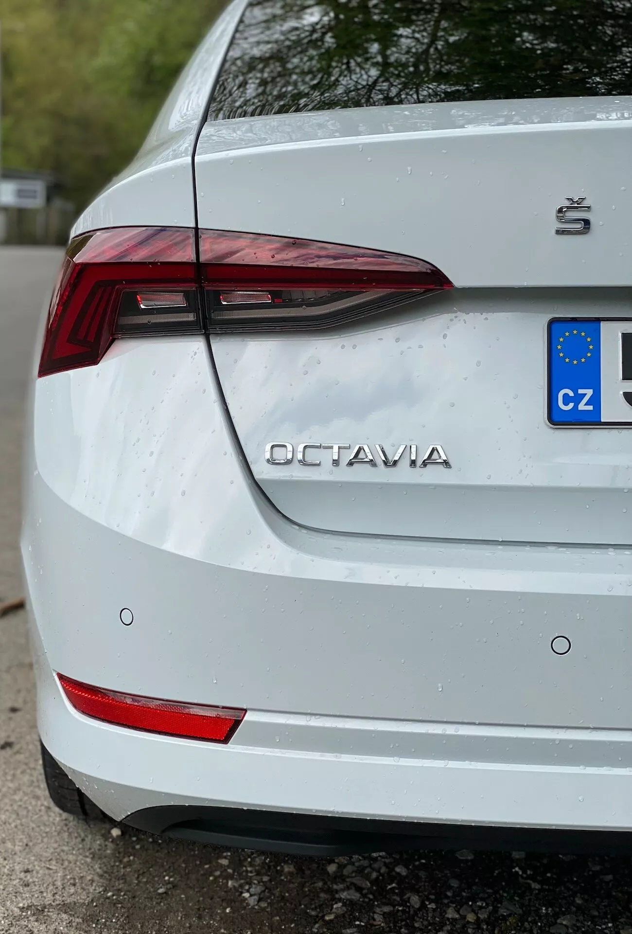 Vue détaillée en gros plan d'un trois-quarts de la partie arrière d'une Škoda Octavia blanche de 2020, mettant l'accent sur le côté droit. L'image met en évidence des caractéristiques importantes comme le feu arrière LED angulaire, les éléments réfléchissants sur le pare-chocs arrière, le nom 'OCTAVIA' et le logo Škoda sur le coffre et la plaque d'immatriculation partielle de l'Union européenne avec la désignation 'CZ'. De fines gouttes d'eau sur la surface indiquent une exposition récente à la pluie ou un lavage, améliorant la fraîcheur de la peinture blanche tout en montrant une légère saleté autour de la plaque d'immatriculation.