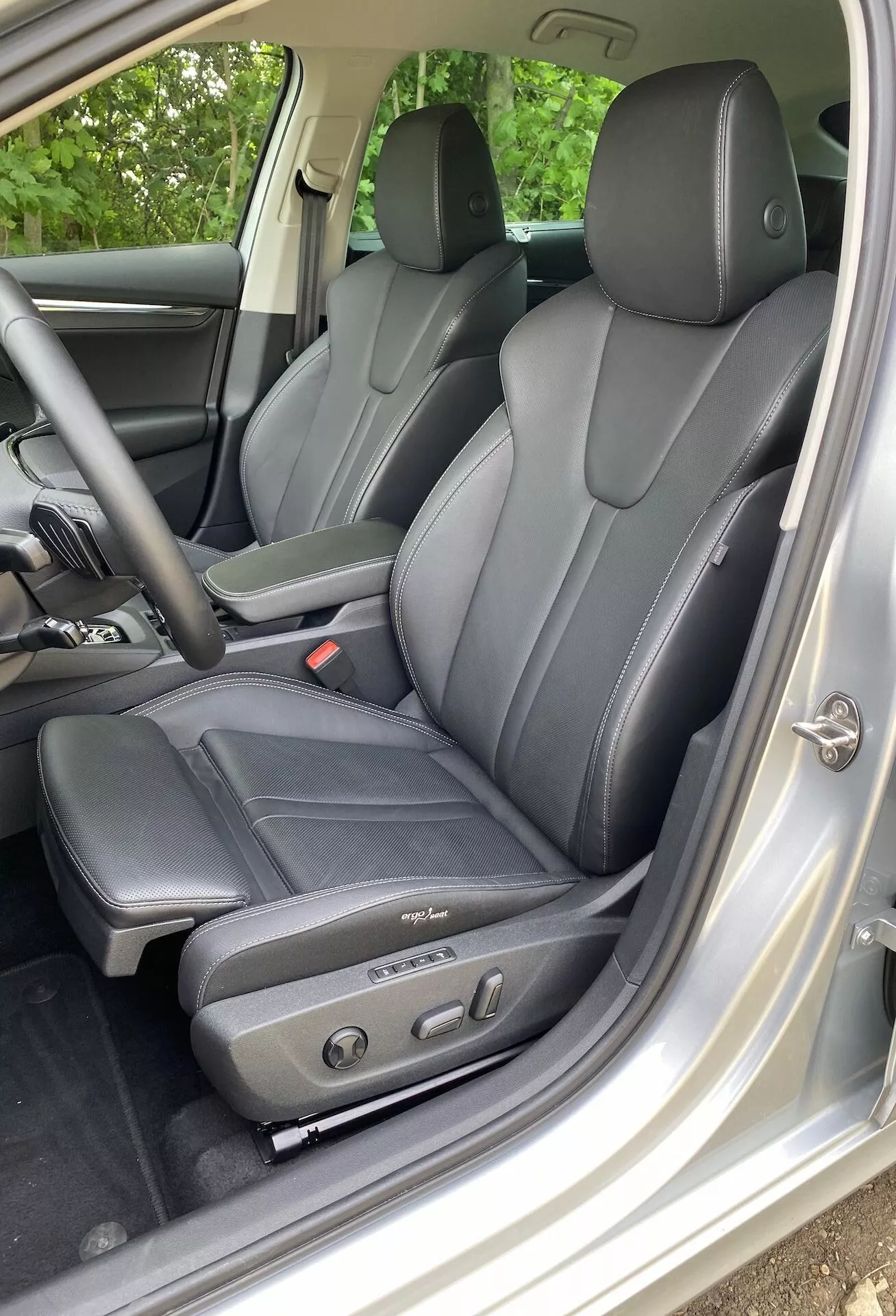 Vue intérieure de la Škoda Octavia limousine 2020 centrée sur la zone du conducteur, avec des sièges en cuir foncé au design ergonomique et des commandes de réglage électrique, vue du côté gauche par la porte du conducteur ouverte. Les appuie-tête visibles pour le conducteur et les passagers arrière indiquent une capacité d'au moins quatre occupants. La finition intérieure argentée ou gris clair contraste avec les sièges, et le panneau de porte comprend des commandes de vitres et de rétroviseurs électriques, mettant en avant le confort et le luxe technologiques.