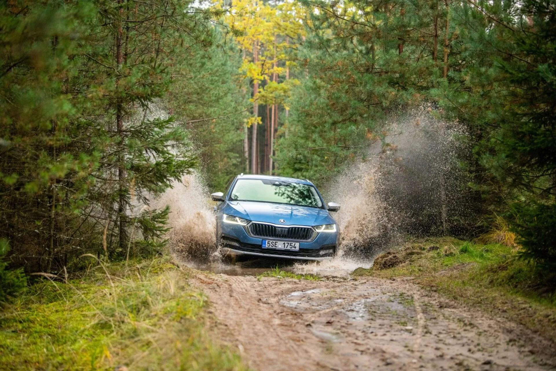 Limousine Skoda Octavia de 2017 traversant une flaque d'eau boueuse dans une zone boisée, mettant en évidence sa création dynamique d'éclaboussures, sa grille avant, ses phares et son capot avec une vue partielle du toit depuis un angle avant droit.